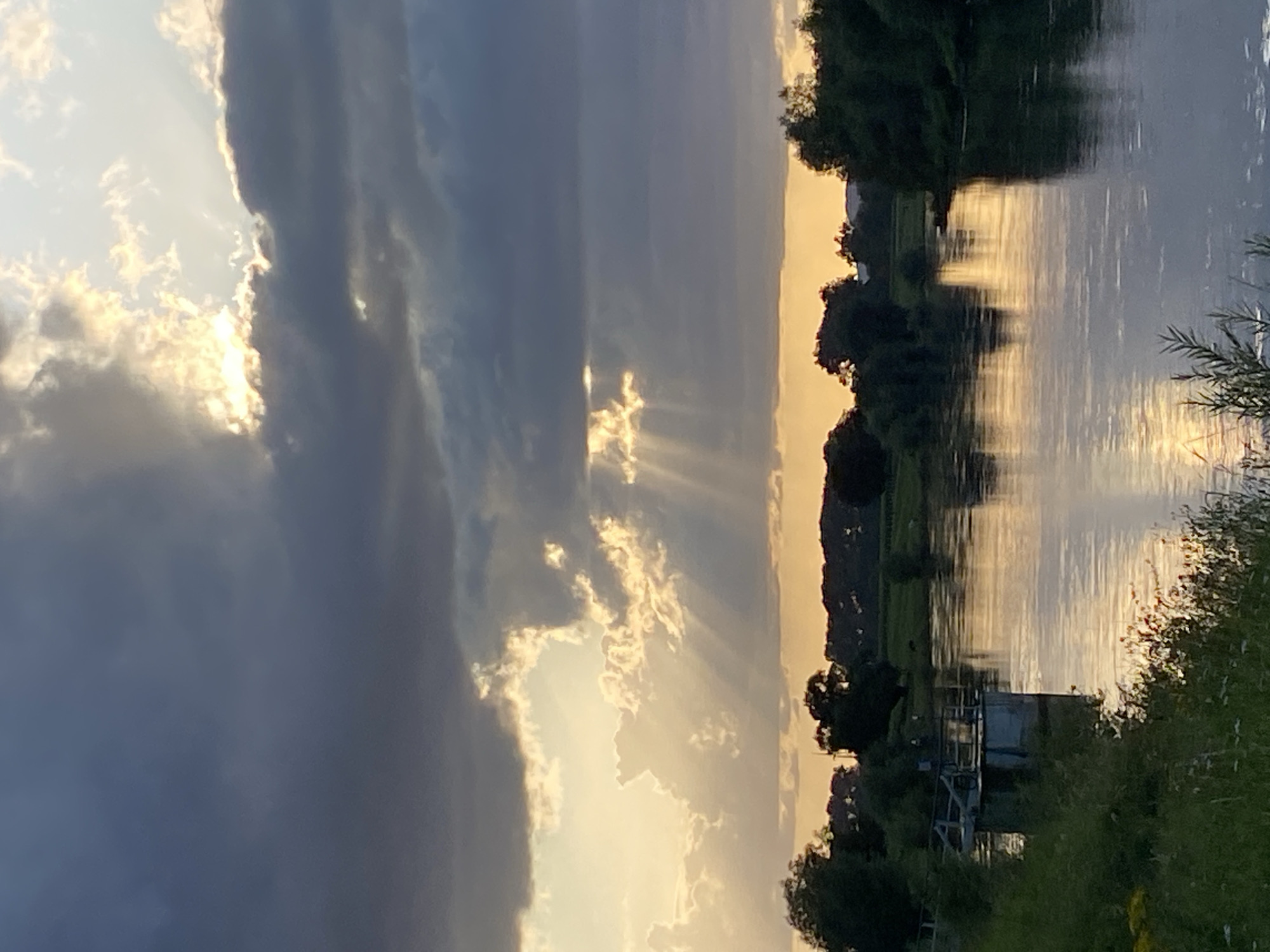The image depicts a wide, tranquil river flowing through a landscape that includes trees and fields, likely at dusk. The sky is dramatic, with dark, heavy clouds partially obscuring the setting sun, which casts rays of light through the breaks. The water reflects the warm glow of the sunset and the dramatic sky. On the left bank of the river, there is a small, industrial-looking structure, possibly a pumping station or part of a lock system, with some vegetation growing around it. The distant shoreline is lined with trees, and beyond them, the faint silhouette of hills can be seen. The overall impression is one of natural beauty and a peaceful, late-day atmosphere. This scene is located on the Weser river in the city of Rinteln, Germany.