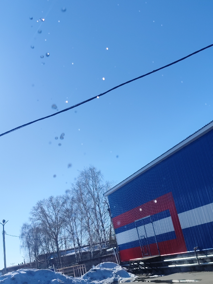 The image captures an outdoor scene in Chaikovsky, Russia. The precise location within the city is not discernible from the visual information. The upper portion of the frame is a clear, bright blue sky, traversed diagonally by a dark power line from the upper left to the middle right. Scattered across the sky and upper left are numerous out-of-focus circular artifacts, consistent with water droplets or dust on the camera lens.
On the right, a large industrial-style building is visible, constructed with blue corrugated metal siding. A distinct rectangular section on its facade is adorned with horizontal stripes of red, white, and blue, containing a dark, closed double door elevated above ground level. This entry point is fronted by a loading dock-like structure with metal railings.
To the left of the building, in the mid-ground, a line of leafless deciduous trees stands against a slightly elevated background. The lower-left foreground displays irregular patches of melting snow and dark puddles on the ground. A black streetlight pole, equipped with two luminaires, is partially visible on the far left. No individuals or specific actions are depicted in the scene.