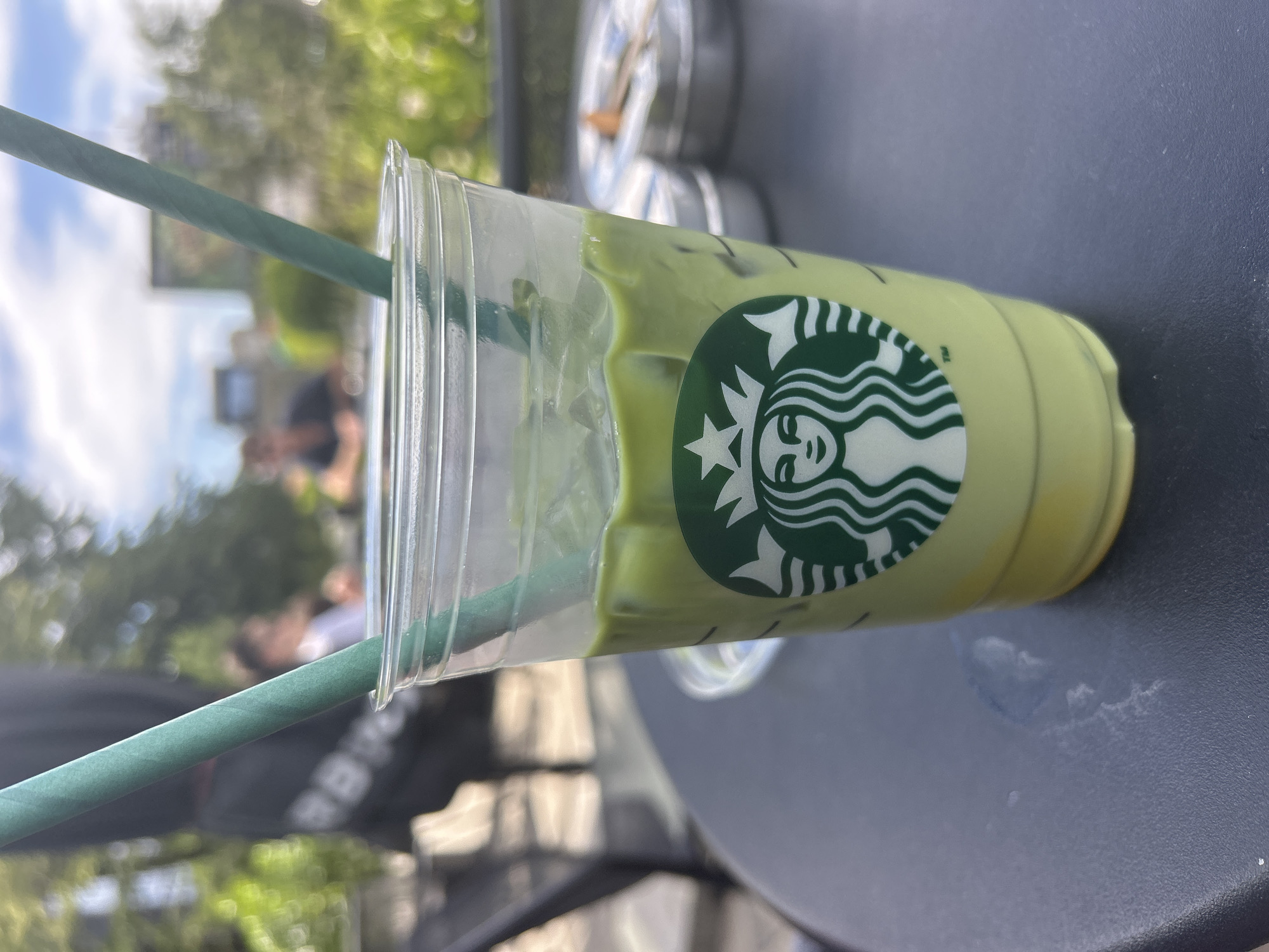 A Starbucks cup with a green matcha latte and a green straw sits on a dark gray surface, likely a table. The cup is partially transparent, revealing layers of ice and the green beverage. The Starbucks logo is prominently displayed on the cup.
In the background, there is a blurred outdoor scene with trees and people, suggesting a public space. The location is a Starbucks, and the outdoor scene appears to be in Sofia, Bulgaria.