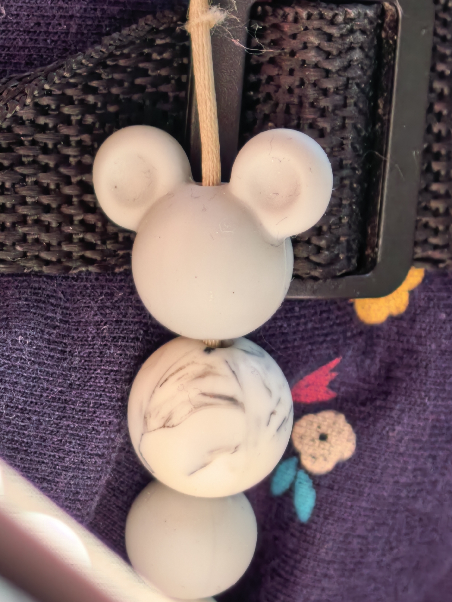 A close-up, overhead view depicts a vertical stack of three decorative beads against a multi-textured background. The top bead is matte white, molded into the recognizable shape of a Mickey Mouse head, featuring a larger central sphere and two smaller, rounded ears. A light tan, rope-like string passes through the top center of this bead and extends upwards. The middle bead is spherical, off-white, and exhibits irregular grey-black marbled patterns. The bottom bead is spherical and matte white.
The background consists of two primary textile surfaces and a dark hardware element. In the upper portion, a dark, tightly woven strap or webbing with a distinct criss-cross pattern is visible, appearing to be black or very dark grey. To the upper right, a black, rectangular buckle or loop component is partially visible, through which the light tan string also passes. Fine dust and fibers are present on both the woven strap and the buckle.
The lower half of the image features a purple fabric, likely knitted, which includes several embroidered elements: a yellow four-petal flower with a darker center, a solid red leaf-like shape, a light beige/tan four-petal flower with a dark center, and two teal-blue leaf-like shapes. The fabric also shows scattered fine dust and fibers. In the bottom-left corner, a blurry, out-of-focus light-colored object, possibly white or pale pink and smooth, intrudes into the frame at a diagonal angle.
The specific location in the city of Sterlitamak, Russia, or any city, cannot be determined from the visual evidence provided in the image.