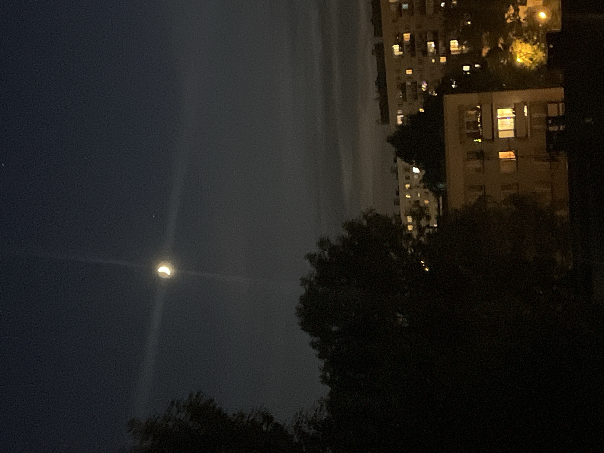 An outdoor nighttime shot captures a crescent moon high in a dark sky above apartment buildings in an urban setting. The moon is positioned in the upper center of the frame, glowing brightly. A faint light streak extends vertically from the moon into the upper sky. Silhouetted trees are visible in the foreground on the left and in the middle ground, obscuring parts of the buildings. The apartment buildings, illuminated by interior lights visible through windows, are spread across the lower portion of the image. Some trees are also visible in the middle ground, interspersed with the buildings. The overall atmosphere is dark and somewhat hazy.