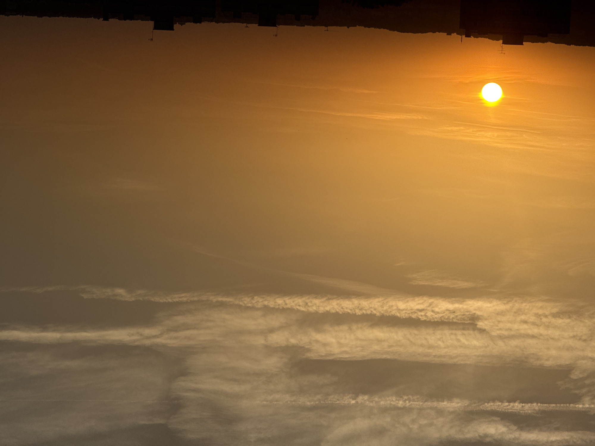 The image depicts a sunset in an urban environment. The sky is a gradient of warm colors, transitioning from bright yellow near the horizon to a muted orange and brown higher up. Wispy, horizontal clouds are scattered across the sky, with some appearing as thin streaks and others as broader bands.
In the lower portion of the image, a silhouetted cityscape is visible against the vibrant sky. The silhouettes include the tops of buildings and some distant hilly terrain. One prominent building in the foreground features an antenna. The sun itself is a bright, white orb positioned in the left half of the frame, casting a warm glow across the scene and creating a stark contrast with the darkening landscape below. The overall impression is one of a serene evening in a city.