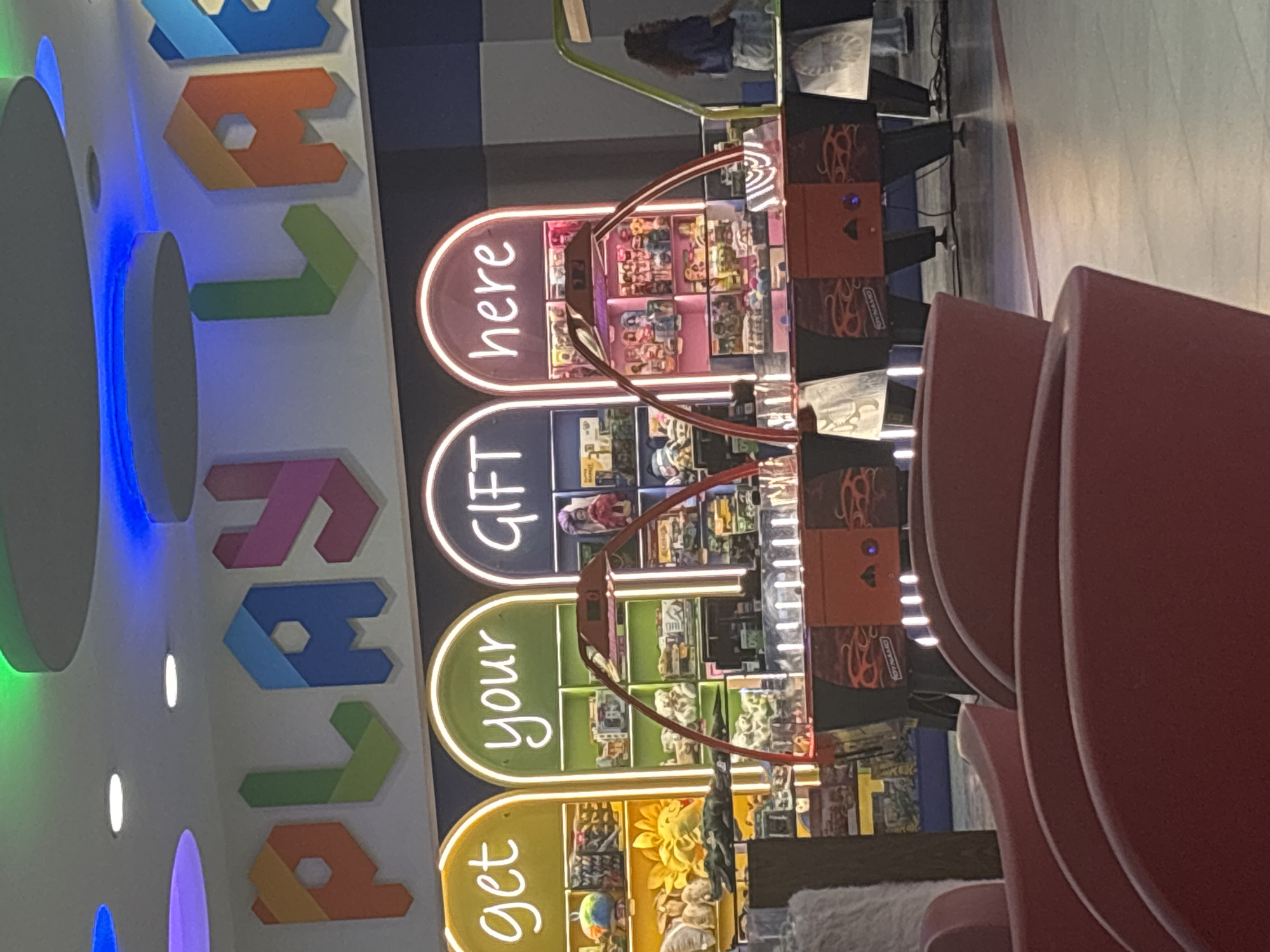 This image captures an indoor entertainment area, likely an arcade or a section within a larger family-oriented venue, located in Yerevan, Armenia. The scene is dominated by bright, artificial lighting.
On the left side of the frame, large, three-dimensional block letters spelling "PLAY" are mounted on a light-colored wall. These letters are rendered in various vibrant colors, including orange, green, blue, and pink, and are internally lit. Blue neon-like strip lighting runs along the top and bottom edges of this wall feature.
To the right of the "PLAY" sign, four vertically stacked, oval-shaped signs with illuminated borders read "get," "your," "GIFT," and "here" from bottom to top. These signs are positioned above and alongside a series of arcade claw machines. Each machine features a transparent case displaying a variety of prizes, primarily plush toys and other small novelty items. The bases of these arcade units are orange with black decorative patterns, and they have visible control panels or monitors. A metallic claw mechanism is visible within the clear casings, ready to retrieve prizes.
In the background, a person with long dark hair is partially visible, seated on what appears to be a chair, reflected faintly in a surface beyond the arcade machines. In the immediate foreground, out of focus, are portions of red, upholstered chairs or seating, indicating a lounge or waiting area for visitors. The overall ambiance is bright and colorful, suggesting a fun and engaging environment.