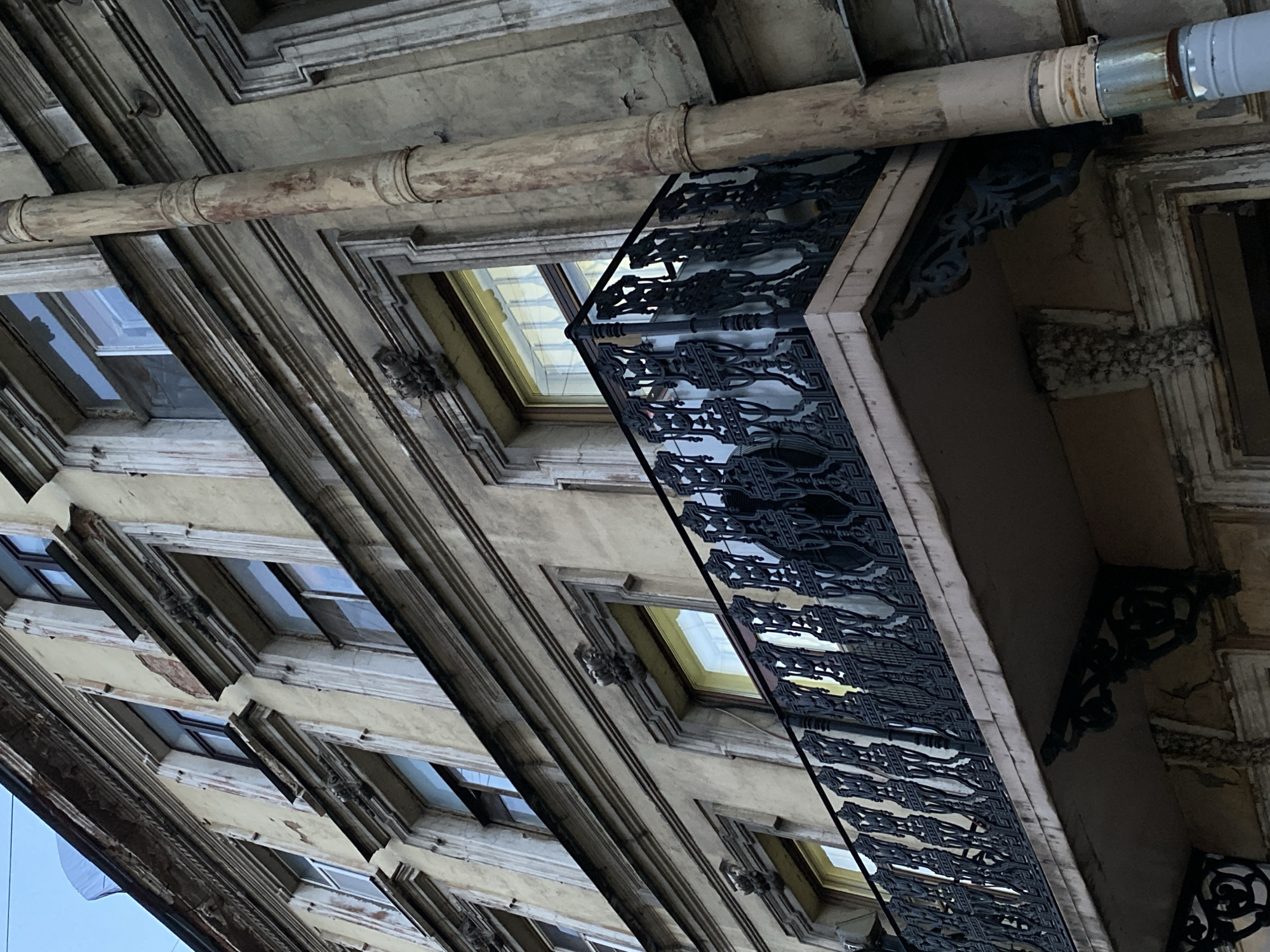 An upward-tilted shot shows the facade of a building in Saint Petersburg, Russia. The building is characterized by aged yellow walls, rectangular windows recessed into the structure, and detailed window frames. An ornate black iron balcony with decorative railings extends from the building. Above the balcony and windows, a horizontal pipe runs parallel to the facade. The sky is faintly visible through the windows and at the bottom left of the frame.