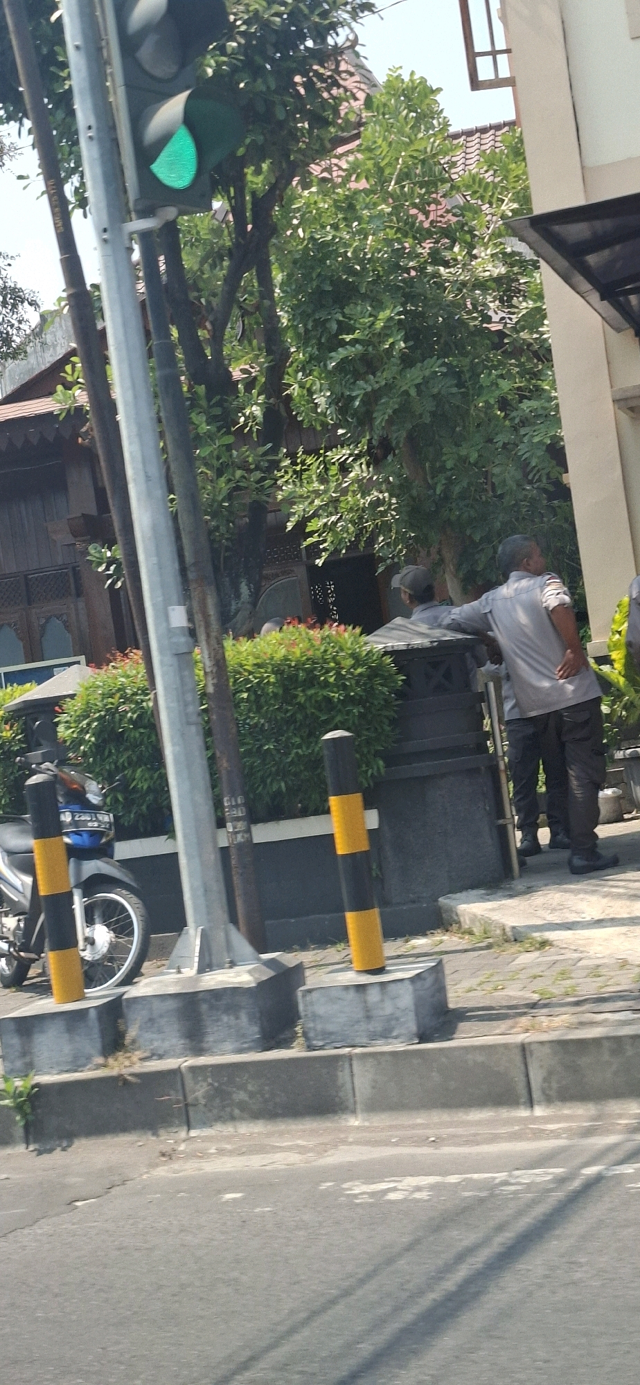 The image depicts a street scene in Surakarta, Indonesia, on a sunny day. A traffic light is visible with a green signal illuminated. In the foreground, a motorcycle is parked. Two men wearing grey uniforms and dark pants stand near a low wall with bushes. One man is leaning against the wall, facing away from the camera, while the other stands beside him, also facing away. A building with a dark-tiled roof and traditional wooden architectural elements is partially visible in the background, surrounded by lush green trees. The street has a concrete curb and sidewalk. Yellow and black striped bollards are present on the sidewalk.