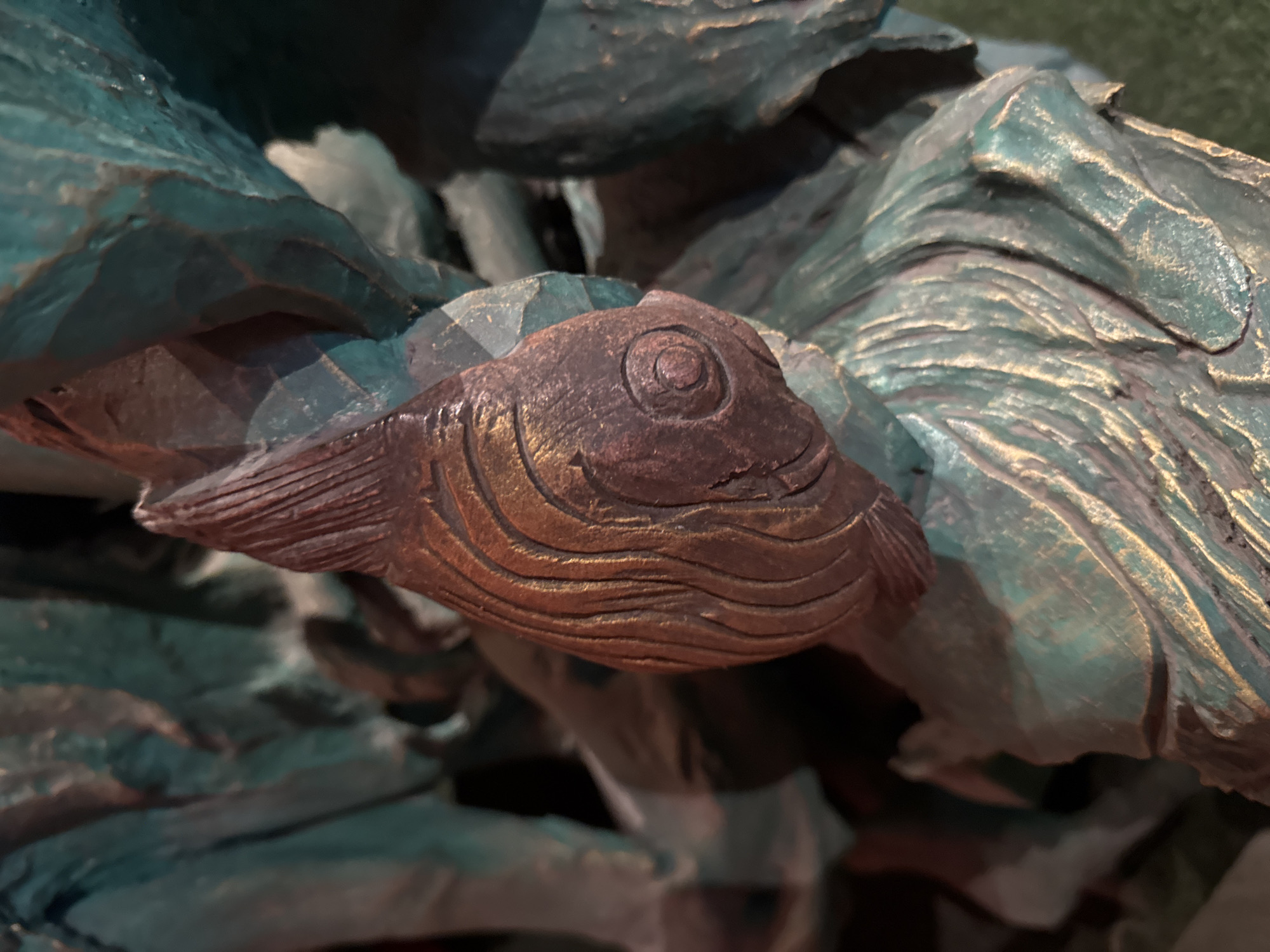 This is a close-up, low-angle shot of a carved wooden sculpture, likely depicting a fish swimming amongst stylized foliage. The sculpture is rendered in a rustic, artistic style with a patina that gives it an aged, weathered appearance.
The main subject is a fish, carved in a deep brown wood. Its head is prominent, featuring a large, circular eye with a concentric circle detail. The body of the fish is textured with several carved wavy lines, suggesting scales or the movement of water. The fins are also intricately carved.
Surrounding the fish are elements that resemble large leaves or petals, carved from a different material or finished with a distinctive greenish-blue patina with golden highlights. These organic shapes form a backdrop and frame for the fish, suggesting an aquatic or naturalistic setting. The textures of these elements are rough and carved, with deep crevices and raised ridges that catch the light.
The lighting is dramatic, with strong highlights and deep shadows that accentuate the three-dimensional form of the sculpture and its textures. This suggests it might be displayed indoors under artificial lighting or outdoors in dappled sunlight. There is no visible text in the image. The overall impression is one of a handcrafted piece of art, evoking a sense of natural beauty and artistic expression. The location context of Kuta, Indonesia, might suggest the sculpture is part of a local art installation or decorative element in a resort or public space, possibly with a theme related to marine life or tropical flora.