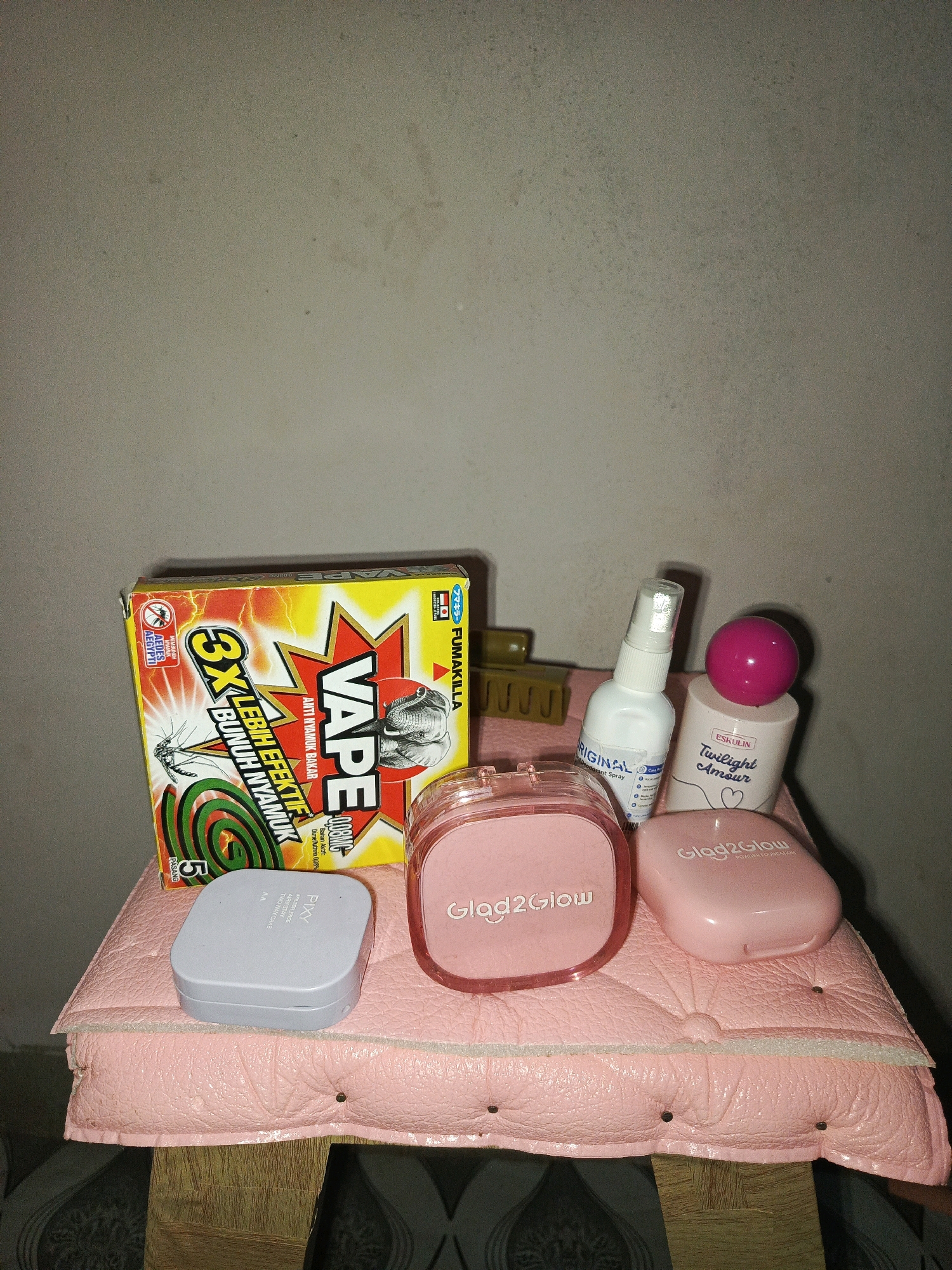 A collection of beauty and personal care products is arranged on a pink, quilted surface, which is resting on a wooden structure. The items include a box of "VAPE" mosquito repellent, a white spray bottle labeled "ORIGINAL" with "Deodorant Spray" below it, a pink-capped cylindrical bottle labeled "ESKULIN Twilight Amour," a light gray square compact with "PIXY" printed on it, and two pink compacts labeled "Glad2Glow" and "Glad2Glow POWDER FOUNDATION." The background is a plain, textured wall.