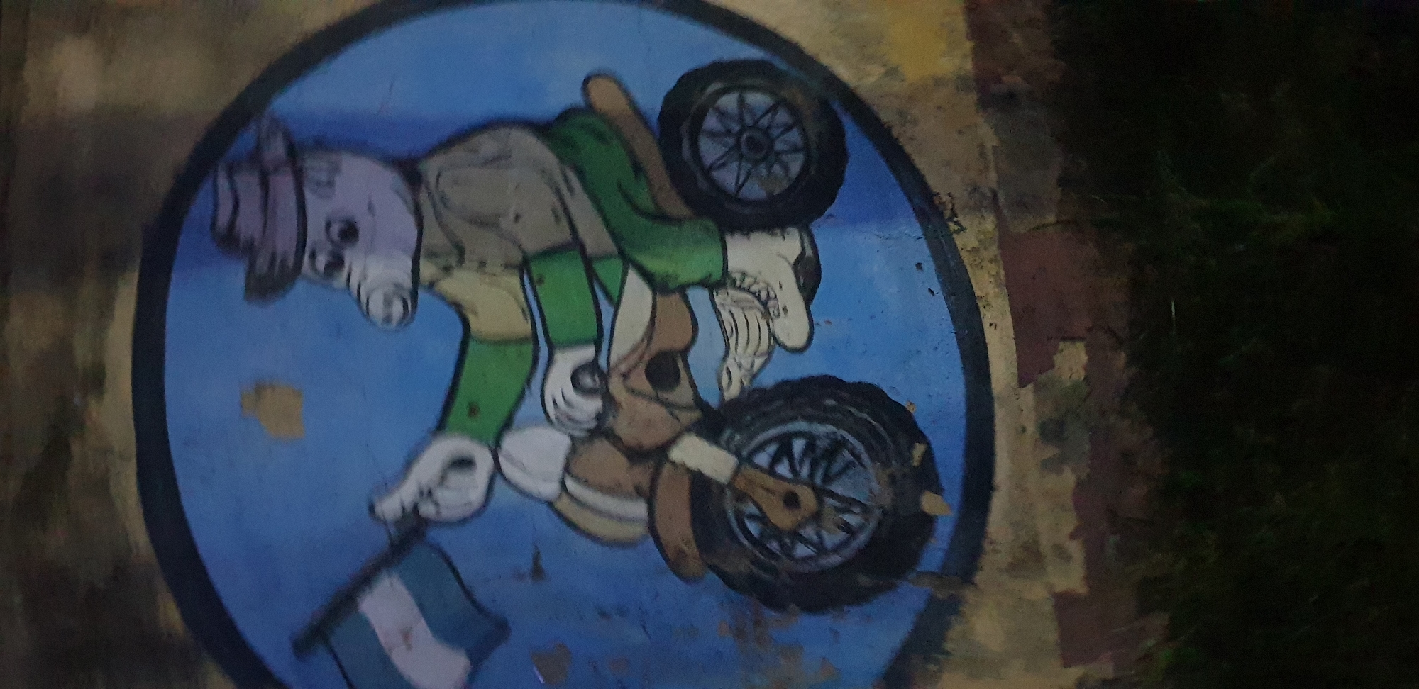 A painting depicts a purple, pig-like creature wearing a hat, green shirt, and green pants, riding a brown motorcycle. The creature is holding a flag with green and white stripes. The painting is on a circular blue background, which is set against a textured, weathered surface. The overall scene appears to be an outdoor mural.