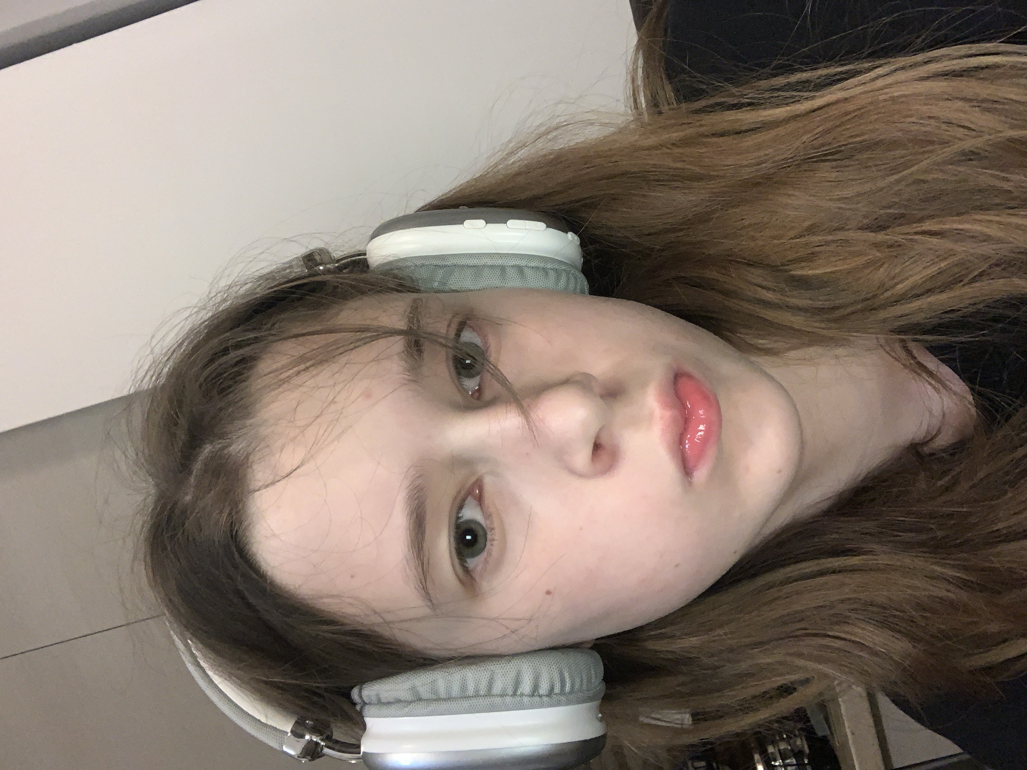 A young person with light skin and brown hair is wearing gray and white headphones. The person is facing the camera, and the image is rotated. The background is a light-colored wall and some dark clothing. No specific location details related to Vozdovac Urban Municipality, Serbia are discernible from the image.