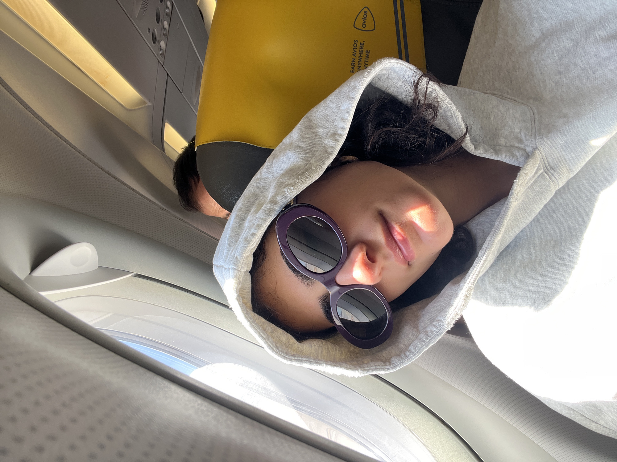 A young woman with dark curly hair, wearing a light grey hoodie and oversized purple sunglasses, is seated by an airplane window, with the sun illuminating her face and hoodie. She is looking towards the camera with her head tilted back slightly. In the background, the yellow headrest of a seat, possibly on a Vueling aircraft due to the "Avios" logo visible, and the back of another passenger's head are discernible. The lighting suggests it is daytime, and the presence of the airplane interior indicates she is in transit. The location is an airplane cabin, not a specific place in Sant Josep de sa Talaia, Spain.