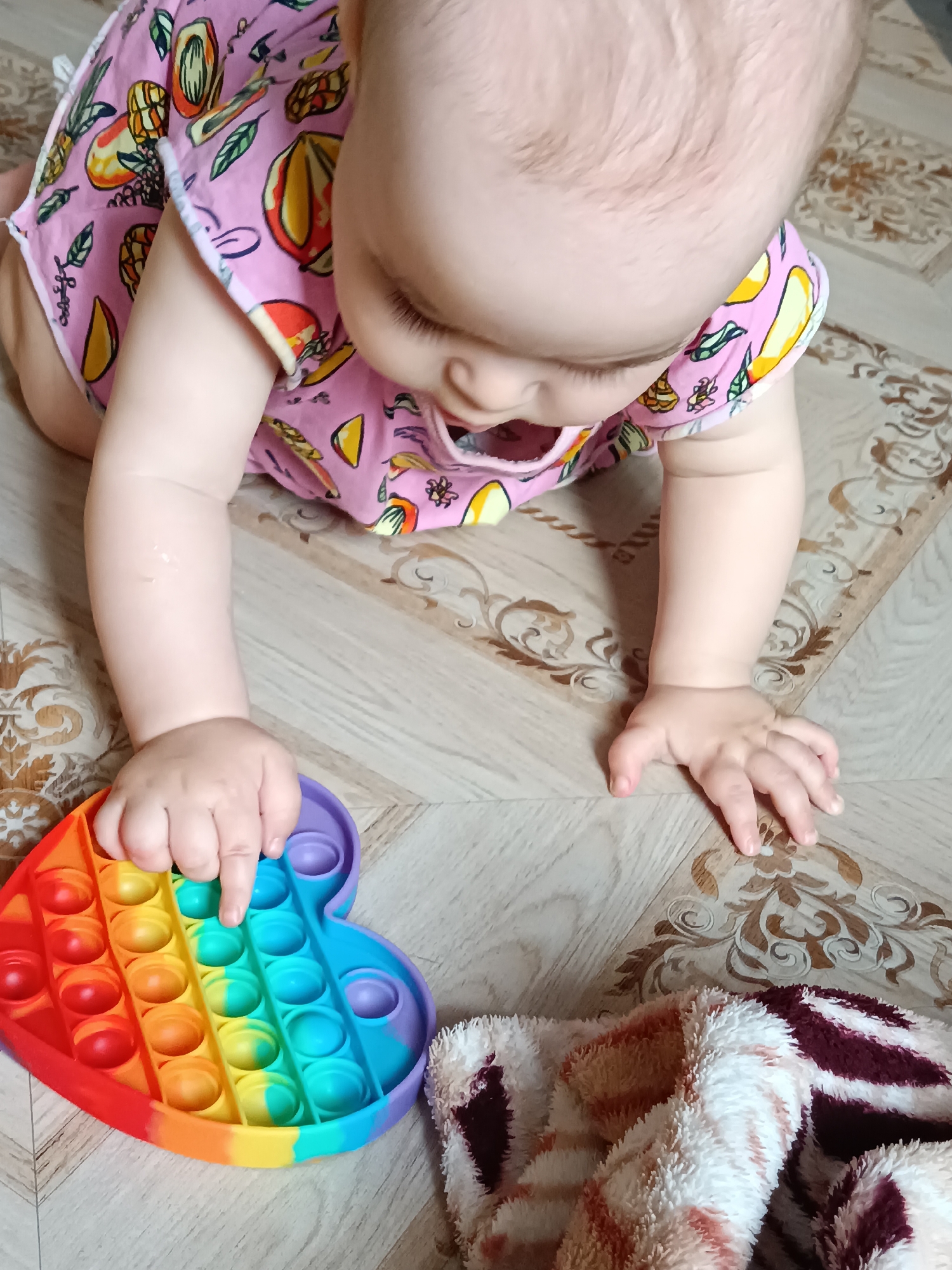The image captures an indoor scene in Noviy, Russia, featuring a young baby or toddler engrossed in play. Viewed from an overhead perspective, the baby is positioned on its hands and knees, looking down with focused attention. It wears a light pink romper or top adorned with a vibrant pattern of fruits like mangoes, lemons, and pineapples, along with green leaves.
The baby's left hand is actively pressing one of the blue bubbles on a rainbow-colored Pop It fidget toy, which is shaped like a heart and rests on the floor. The toy displays a gradient of colors from red, orange, and yellow on the left to green and blue on the right. The baby's right hand is open and resting on the floor beside it.
The floor beneath is light-toned, appearing to be wood or tile, and features decorative brown and beige swirling patterns in sections. In the lower right portion of the frame, a fluffy blanket or textile, striped in white, orange, and dark red, is partially visible. The indoor lighting suggests it is daytime. The baby appears calm and engaged in sensory exploration with the toy. No visible text is present in the image.