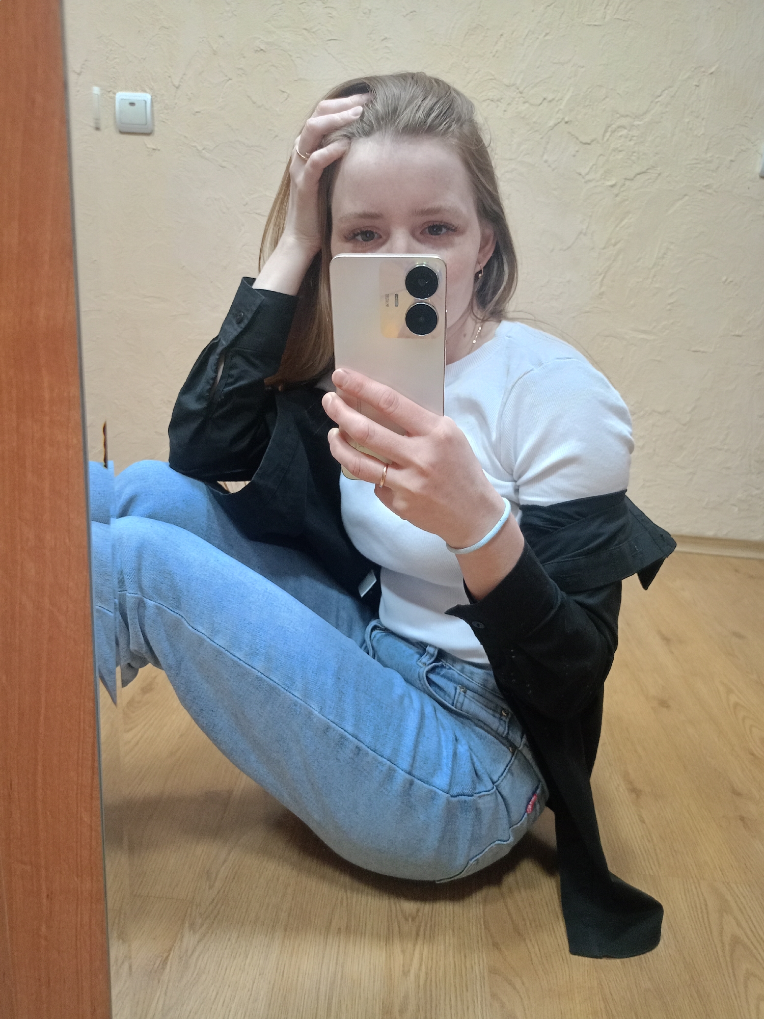 A young woman sits on the floor in front of a mirror, taking a selfie with her cell phone. She wears a white t-shirt, light blue jeans, and a black jacket draped over her shoulders. Her left hand is on top of her head, and she is looking at the phone. The background shows a wall painted in a light beige tone, a light switch, and a wooden floor. The location appears to be indoors.