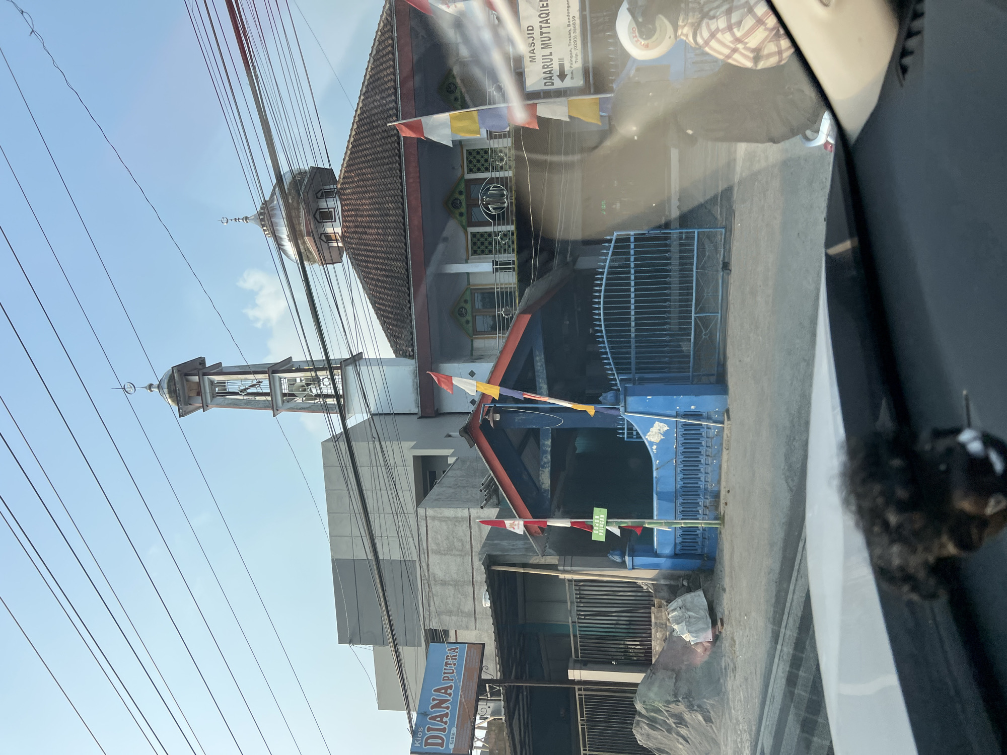 The image captures a street view in Tonoboyo, Indonesia, dominated by a mosque and adjacent buildings. The mosque features a prominent dome and a tall minaret, both topped with crescent moon symbols. In front of the mosque, a blue gate and fence enclose a courtyard. Several flags, including Indonesian flags and white and yellow banners, are displayed. A sign identifies the mosque as "MASJID DAARUL MUTTAQIEN". To the left of the mosque, a building with a sign that reads "Kios DIANA PUTRA SEDIA" is visible. Overhead, a tangle of electrical wires crisscrosses the sky. The ground in front of the buildings appears to be a paved road. A person on a motorcycle wearing a helmet and checkered shirt is partially visible on the right side of the frame, suggesting ongoing activity in the area. The overall scene conveys a typical urban environment with religious and commercial establishments coexisting.