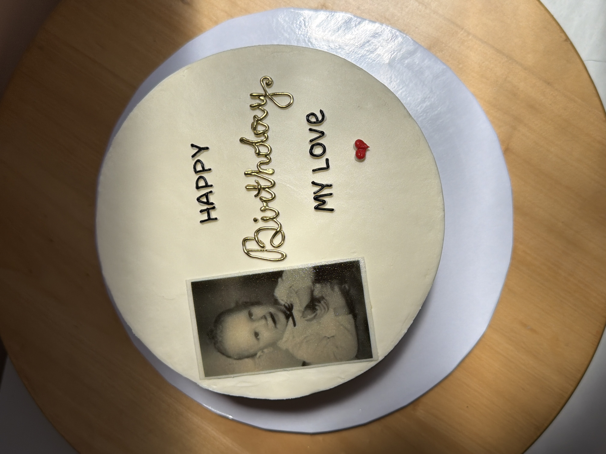 A photograph shows an overhead view of a round cake resting on a white cake board, which in turn is placed on a wooden turntable. The cake has a smooth, white frosting. The words "HAPPY" and "MY LOVE" are written in black on the cake, and the word "Birthday" is written in a cursive gold color. There are two small red hearts made of frosting near the words "MY LOVE." A black and white photograph of a baby is placed on the cake near the bottom. The wooden turntable has a light brown wood grain. The location is not discernable from the image.