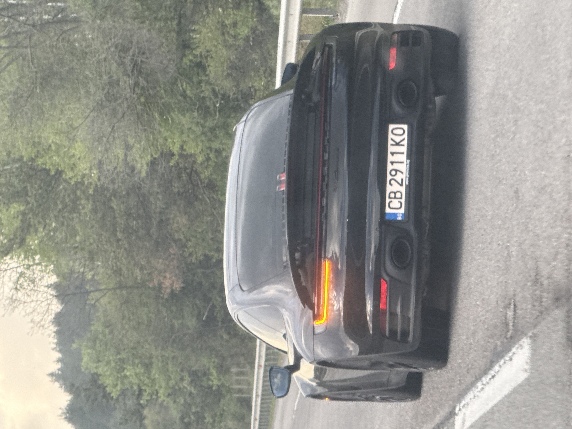 A black Porsche 911 is on a highway, oriented away from the camera. The Bulgarian license plate reads "CB 2911 KO". The location is presumably near Rajana, Bulgaria, due to the license plate originating from Sofia, Bulgaria. There are trees visible on the left side of the highway. A road barrier is on the left side of the road. There is a white lane marking on the road surface. The right turn indicator is illuminated.