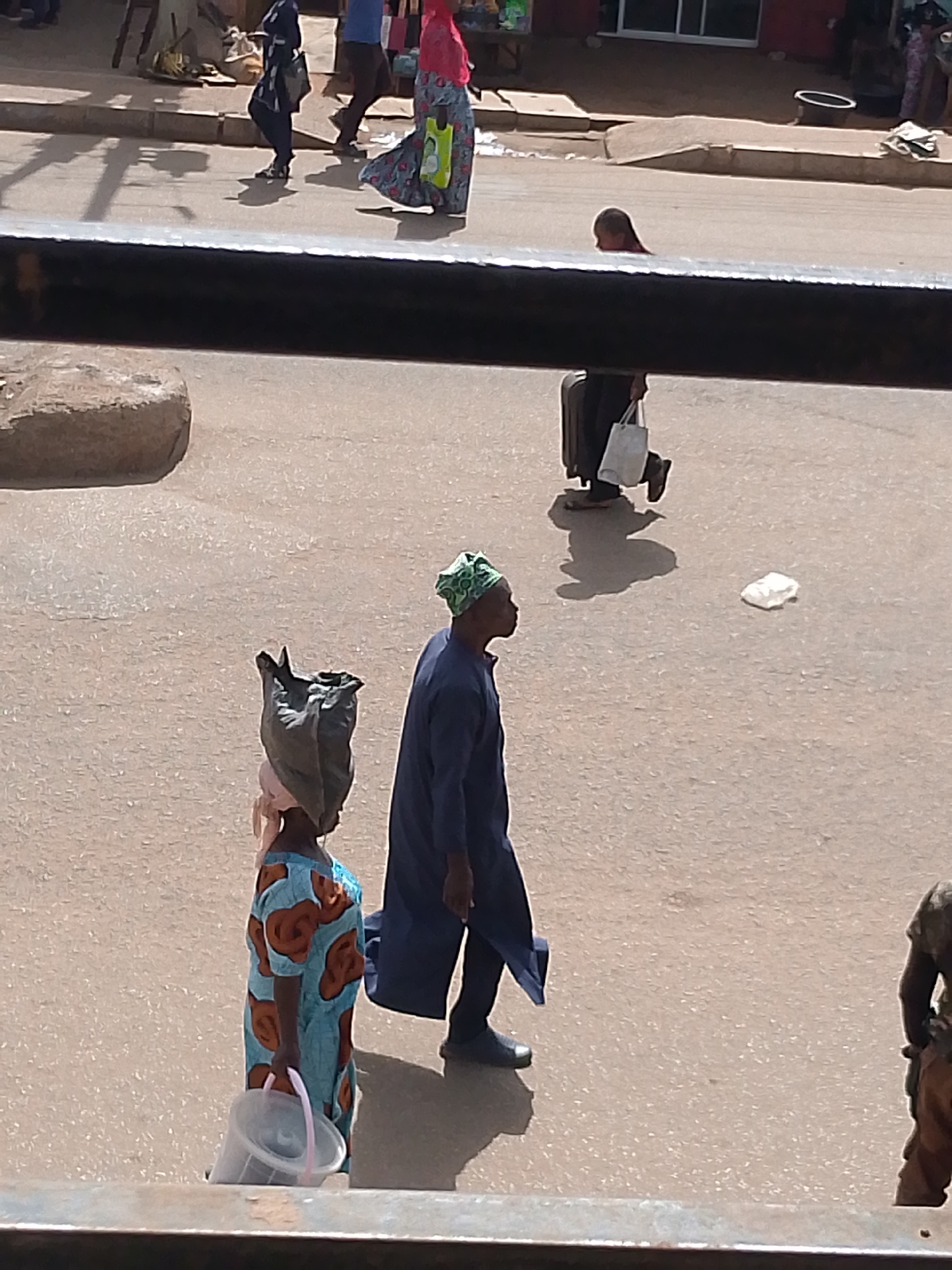 This media file captures a street scene in Bukuru, Nigeria, likely during the daytime given the shadows. Several individuals are walking on a paved street. In the foreground, a person wearing a blue and orange patterned dress carries a large, dark bag on their head and a clear plastic bucket in their right hand. To their right, a person in a dark blue robe and a green head covering walks by, facing left. Further in the background, other figures are visible, including one person pushing a suitcase and carrying a white bag, and another person in a pink top and patterned skirt. The ground appears to be dusty, and the setting suggests an outdoor market or a busy thoroughfare. There are no visible text elements in the image.