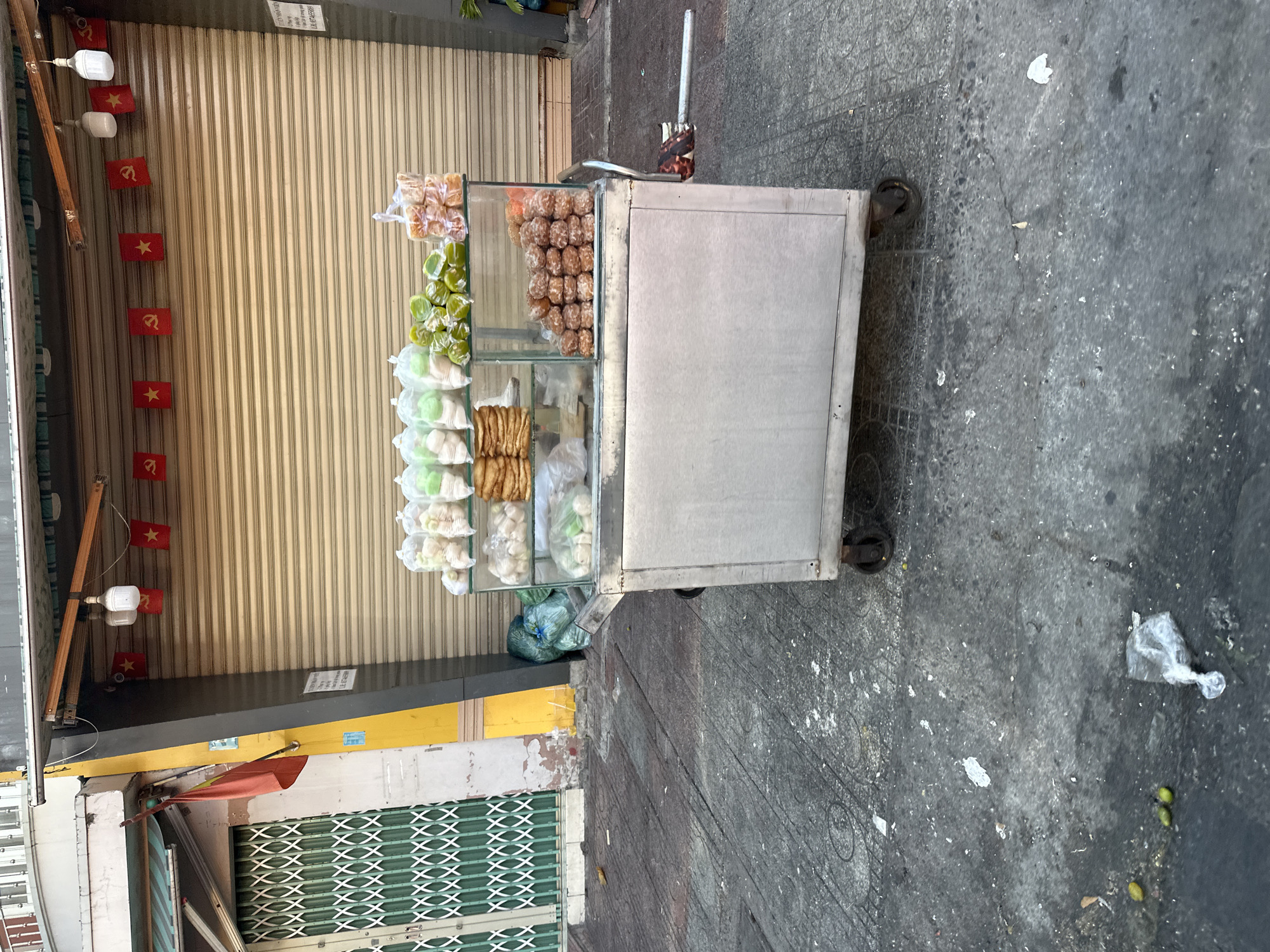 The image depicts a street scene in Bac Nha Trang Ward, Vietnam. In the foreground, a food cart made of stainless steel with a glass display case is parked on a concrete sidewalk. The cart contains an assortment of food items, including what appear to be pastries or donuts, and some green produce. Bags of food are stacked on shelves within the display case. The cart has wheels and a handle, suggesting it is mobile.
Behind the food cart, a large, beige, corrugated metal shuttered storefront is visible. Red rectangular flags with yellow stars are strung across the top of the shutter. The flags appear to be patriotic or political symbols. To the left of the storefront, a building with a green metal gate and an orange flag hanging from a pole is seen. The sidewalk is gray and shows signs of wear and tear, with scattered debris, including plastic bags and what appear to be fallen fruit. The overall environment suggests an urban setting, possibly in the morning or evening given the lighting. There are no people visible in the image.