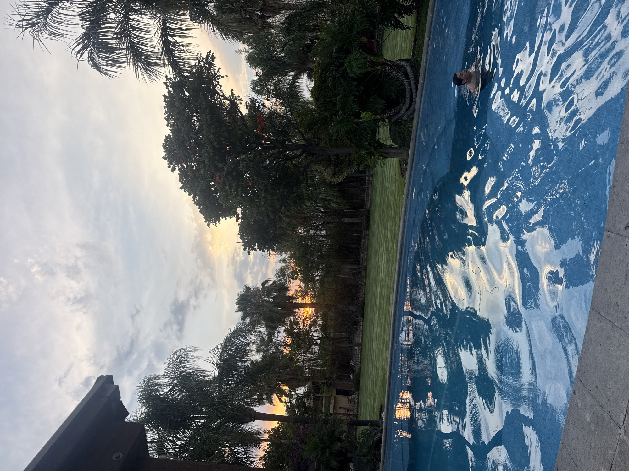 A landscape photograph taken in Santa Ana Yancuitlalpan (Chilhuacán), Mexico. The image shows a swimming pool filled with rippling water, reflecting the sky and trees. A person's head is visible in the water. Trees, including palm trees, surround the pool and a patch of green grass. The sky is visible in the background, with light suggesting it could be near sunrise or sunset. The edge of a building is visible in the bottom left corner.