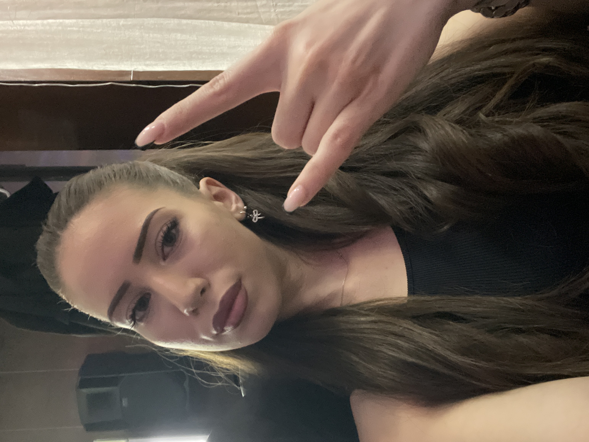 A woman with long brown hair is posing for a selfie. She is wearing a black top. Her right hand is in the foreground, making a "rock on" hand gesture. The background is blurry, showing a window and a dark-colored object that might be a piece of furniture. There is no information available to identify a location in the city of Somovit, Bulgaria.