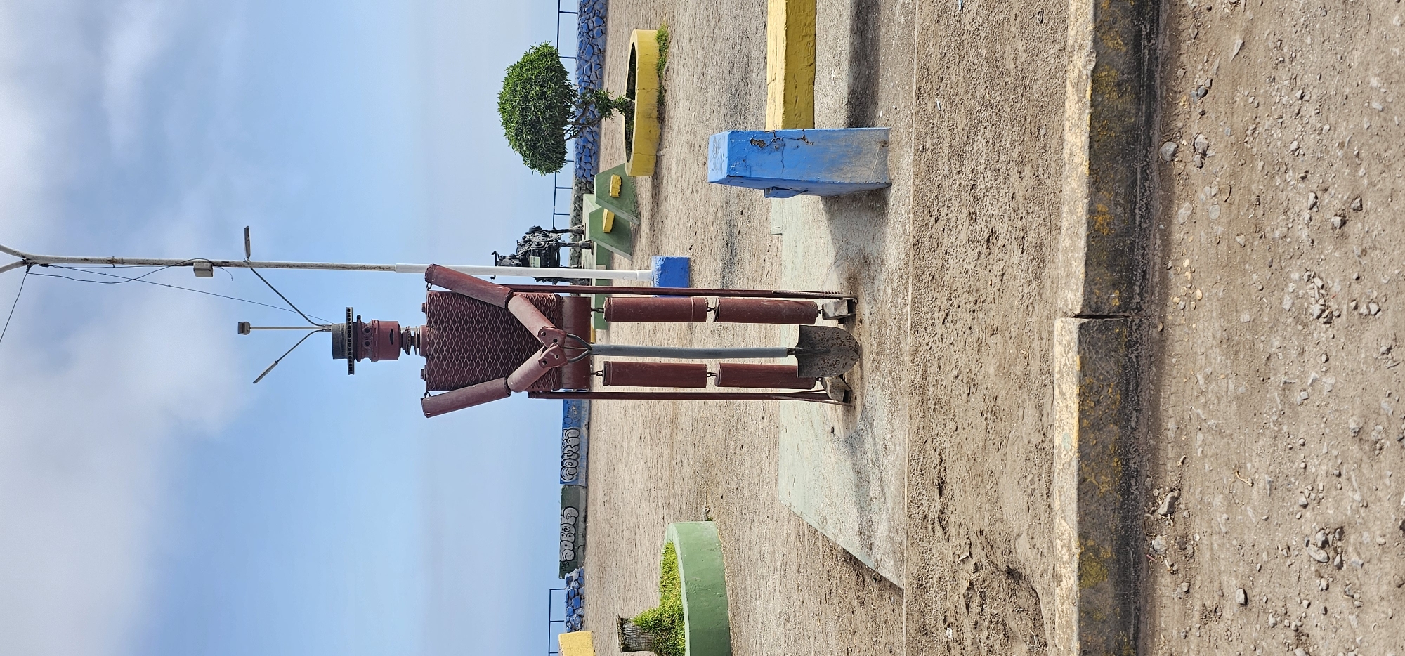 The image shows a metal sculpture of a person in Alto Hospicio, Chile. The sculpture is made of red metal pipes and a diamond-shaped mesh for the torso. It stands on a concrete base. The sculpture is adjacent to a light pole with visible wiring. The background includes a dirt-covered area with various colorful, rectangular, concrete structures and a small tree. In the distance, graffiti can be seen on a low wall. The sky is blue with some cloud cover.