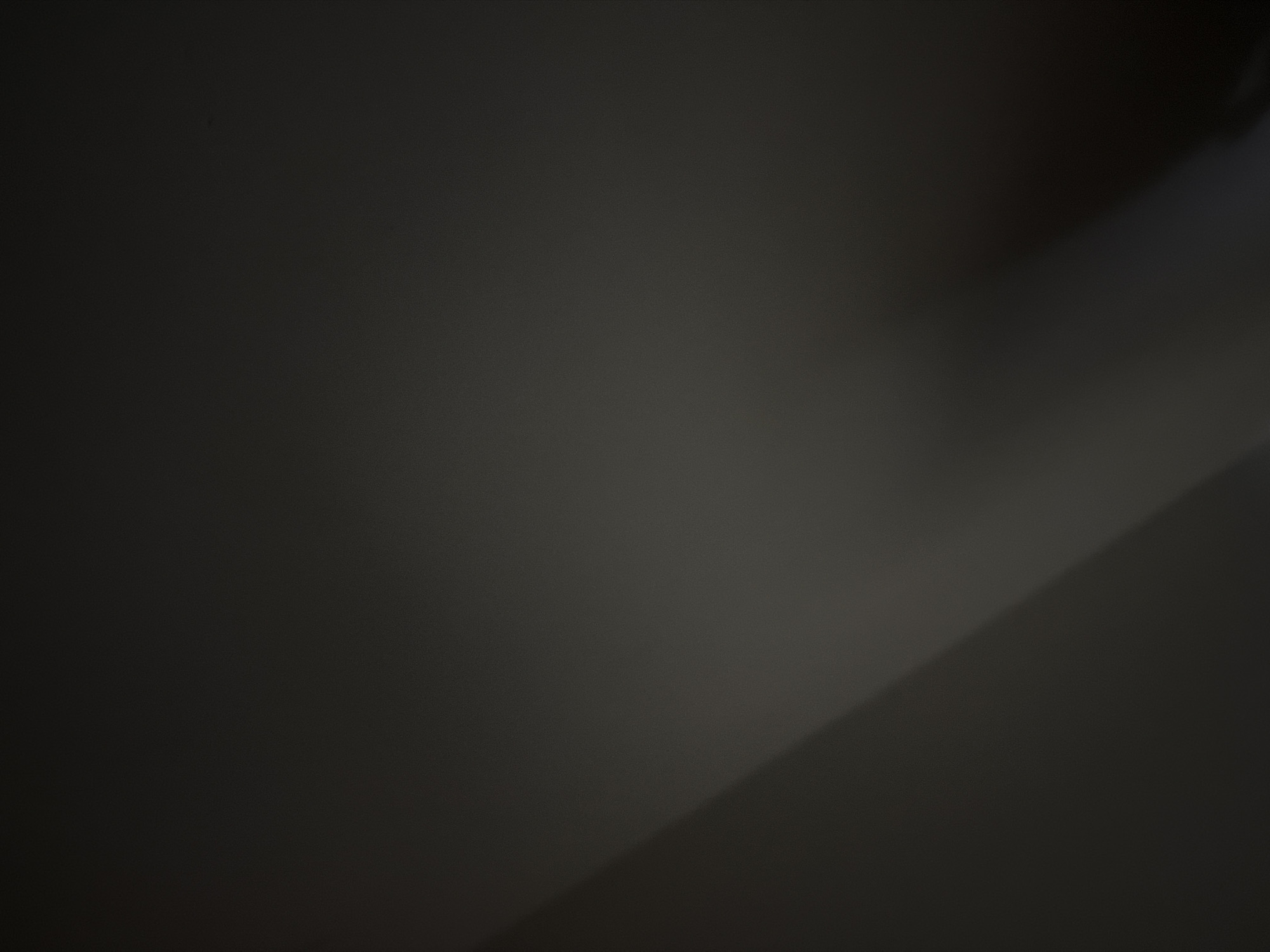 The image is a dimly lit, abstract scene dominated by shades of gray and black. A diagonal line of lighter gray cuts across the upper left portion of the frame, suggesting a shadow or a transition between surfaces. The rest of the image is a dark, textured expanse. There are no discernible people, objects, or environmental features that would indicate a specific location within The Hague or any other city. The overall impression is one of low light and minimal detail.