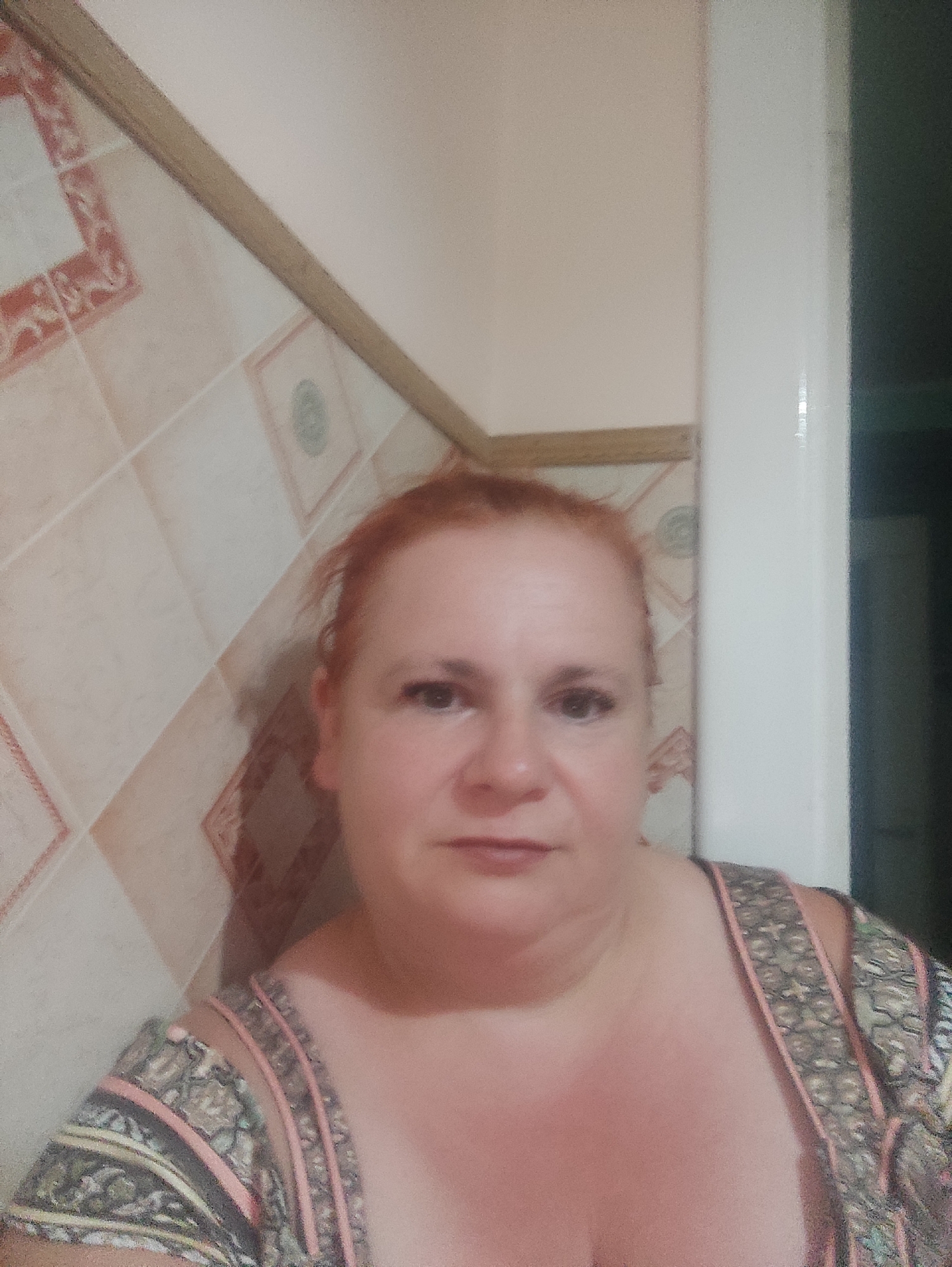 The image shows a selfie of a woman with reddish-blonde hair, taken indoors. She is wearing a patterned top with pink and grey accents. The background consists of a tiled wall with a wooden trim and a plain, light-colored wall. To the right, a white doorframe is visible, with a dark area beyond it. There is no indication of the city of Chernivtsi, Ukraine, in the image itself.