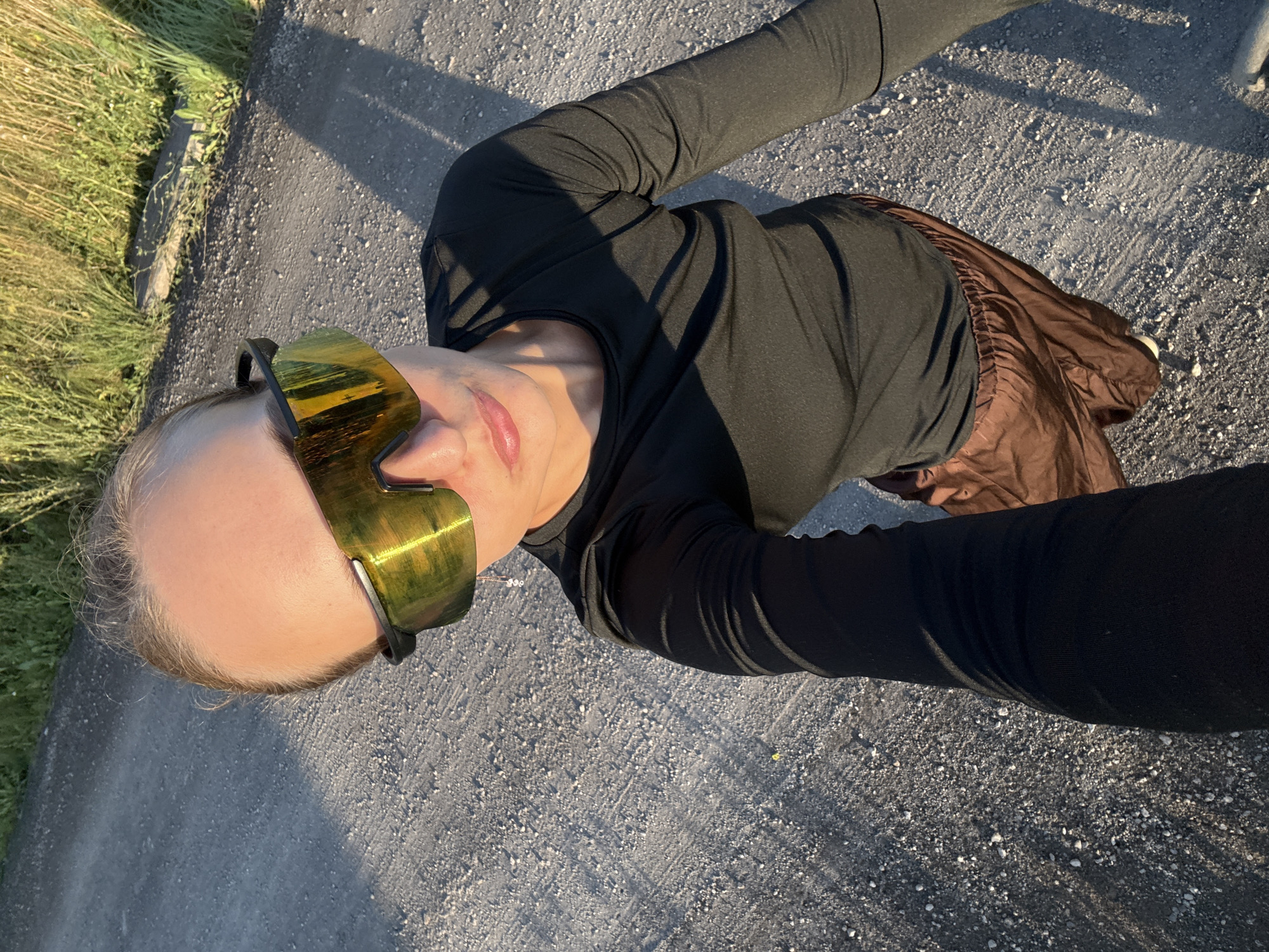 A person is lying down outdoors, wearing a black long-sleeved shirt and brown athletic shorts. They have on large, mirrored wrap-around sunglasses with yellow lenses. Their face is partially visible, and they appear to be looking up towards the camera. The scene is set on a gravel surface, possibly a path or road, with some green grass and foliage to the left. Shadows from the person and possibly nearby objects are cast on the ground, indicating it is daytime, likely late afternoon or early evening due to the warm lighting. The overall mood is relaxed and casual. No text is visible in the image. The location is identified as Podshivalovo, Russia.