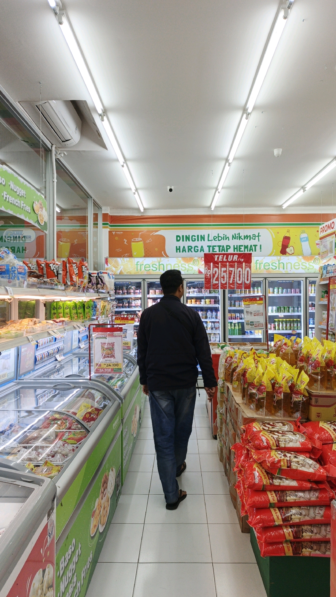An indoor shot captures a man walking away from the camera down the aisle of a convenience store in Parigi, Indonesia. The man is wearing a dark blue jacket and blue jeans. The store is brightly lit by long fluorescent lights on the ceiling, and features rows of refrigerated display cases on the left and shelves stocked with products on the right. Promotional banners and signs are visible on the walls and hanging above the shelves. The floor is tiled in a white grid pattern.