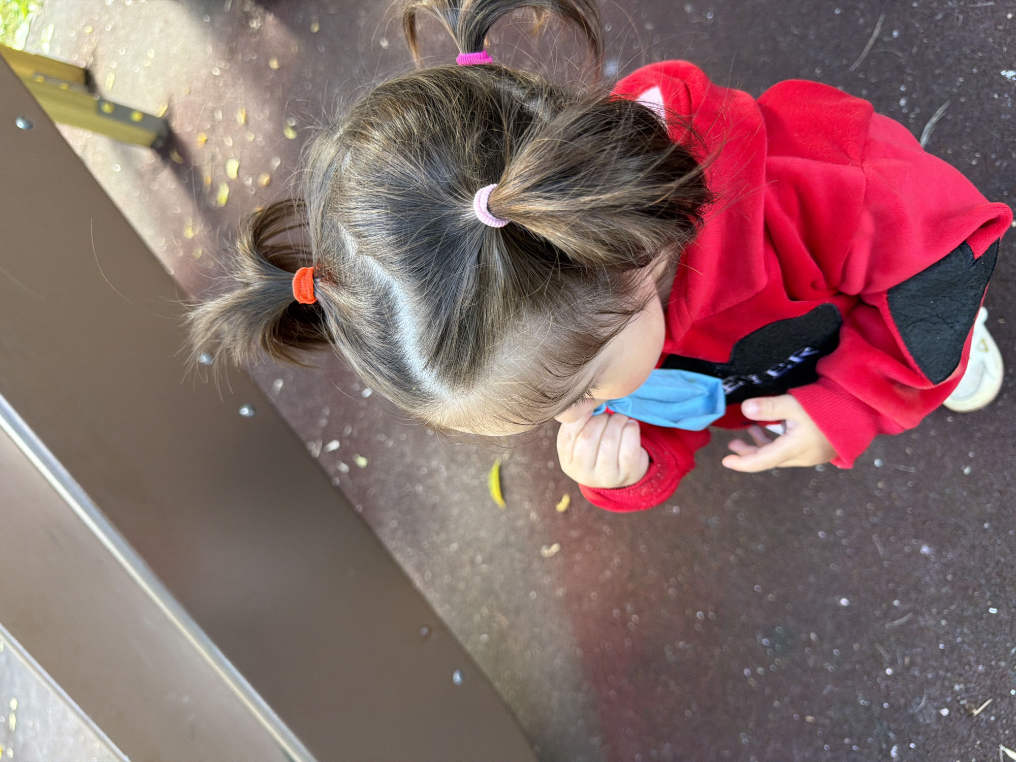 A high-angle, close-up shot shows the back of a young child with dark hair pulled into multiple pigtails, adorned with red, pink, and another pink hair tie. The child is wearing a red hooded jacket with black accents. In their hands, the child holds a blue object, possibly a face mask, near their mouth. The child stands on a dark, textured surface, possibly playground ground, with scattered light-colored debris and some yellow leaves. A brown structure, likely part of a playground slide, is visible in the upper left portion of the frame, with a yellow support post and metal fixtures visible in the upper right. The lighting suggests an outdoor setting on a sunny day, with dappled light and shadows present. The scene is set in a park or playground, which could be located in Moscow, Russia.