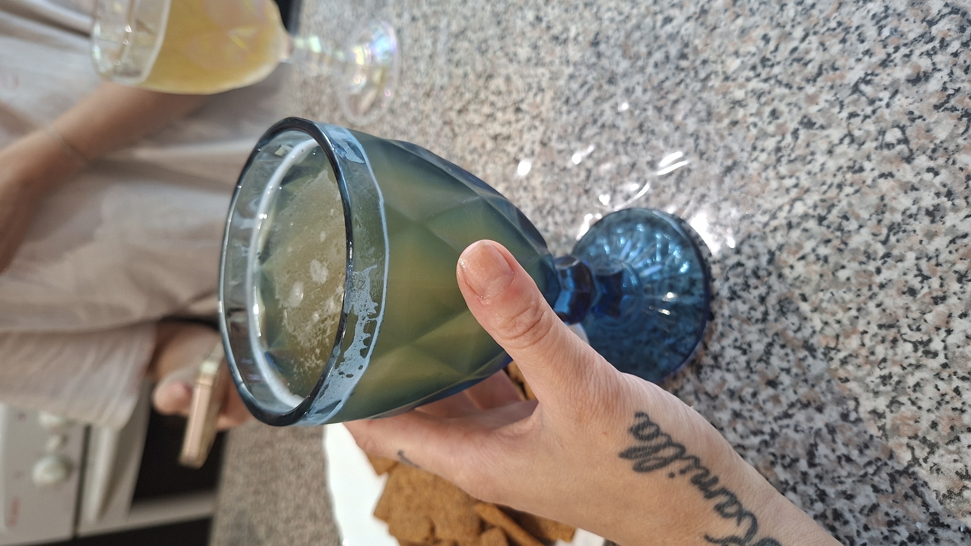 A hand with a tattoo reading "Kamilla" on the forearm is holding a blue glass goblet filled with a light-colored beverage, possibly beer or a cocktail, topped with foam. In the background, a person wearing a white t-shirt is holding a mobile phone. Another glass goblet with a similar beverage is visible to the right. A plate of crackers is positioned to the left of the blue goblet. The scene appears to be set on a speckled granite countertop, likely in a kitchen or dining area. The city of Myadzyel, Belarus, is not depicted in this image.