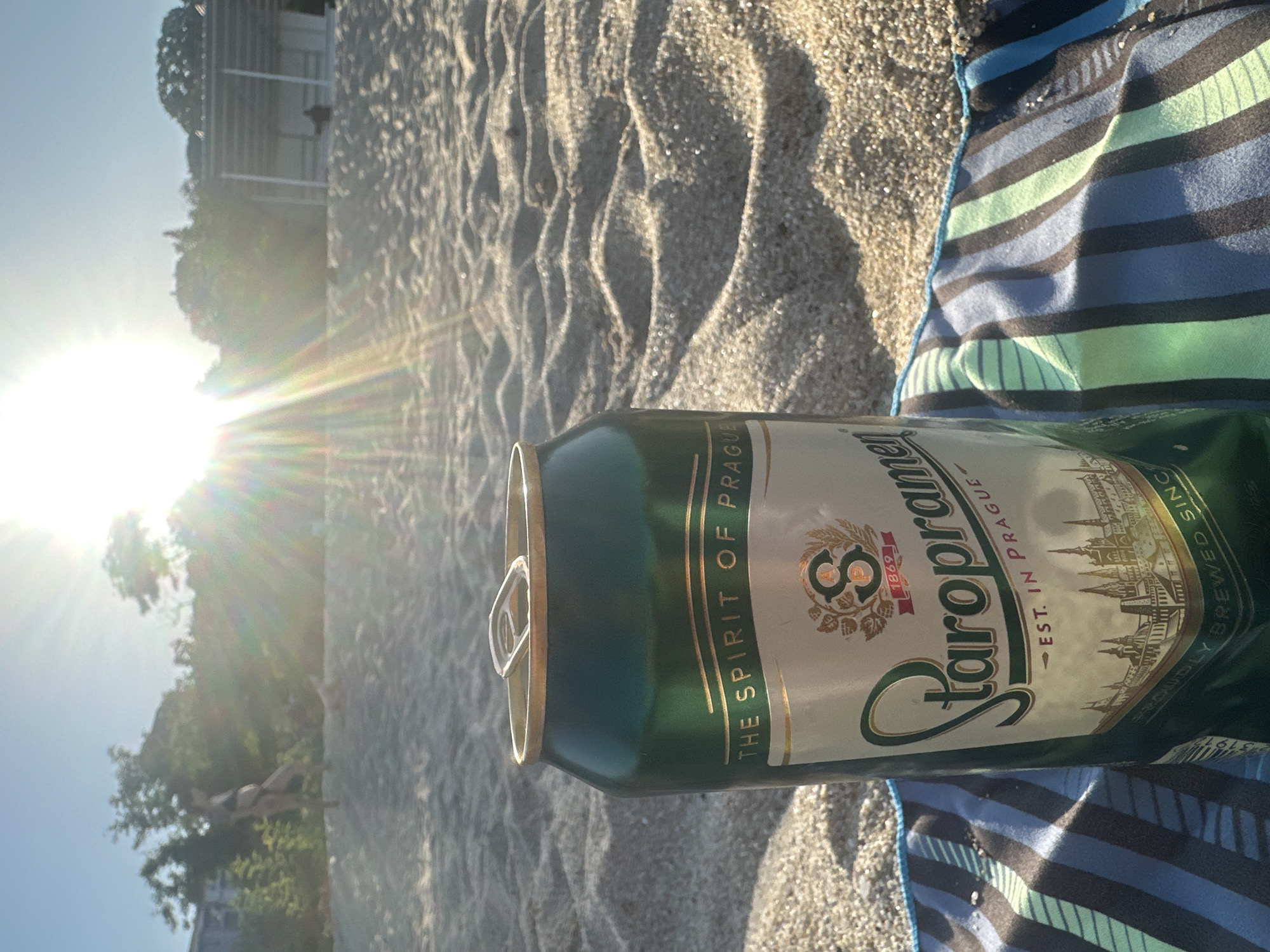 A can of Staropramen beer sits on a striped beach towel on a sandy beach. The can is green with a silver label that reads "THE SPIRIT OF PRAGUE" and "STAROPRAMEN EST. IN PRAGUE". In the background, the sun shines brightly through the trees, casting lens flare on the image. A person in a bikini is visible in the distance on the left side of the frame, and a building or structure is visible in the distance on the right. The sand is rippled, suggesting it has been walked on or affected by wind or waves. The lighting suggests it is either sunrise or sunset.