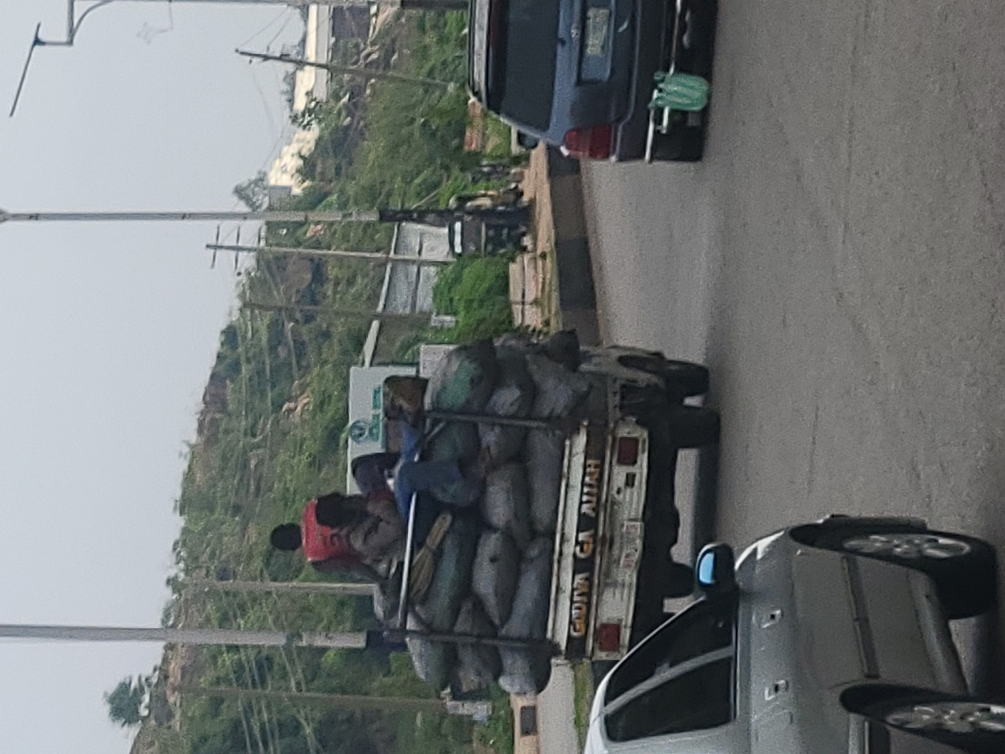 The image captures a street scene in Gura Top, Nigeria, on a cloudy day. In the foreground, the left side of a silver sedan is visible. The main focus is a flatbed truck loaded with numerous large, dark-colored sacks, possibly charcoal or agricultural produce. Three individuals are situated on top of the sacks. One person in the rear is wearing a red vest and appears to be lying down or resting. Two other individuals are seated or leaning against the sacks. The truck has "GADIYA GA ALLAH" inscribed on its rear. Behind the truck, an SUV, also dark-colored, is traveling in the same direction. The environment suggests an urban or semi-urban setting, with a paved road, a curb, and a hillside with sparse vegetation and some structures in the background. Electrical poles and wires are also visible.