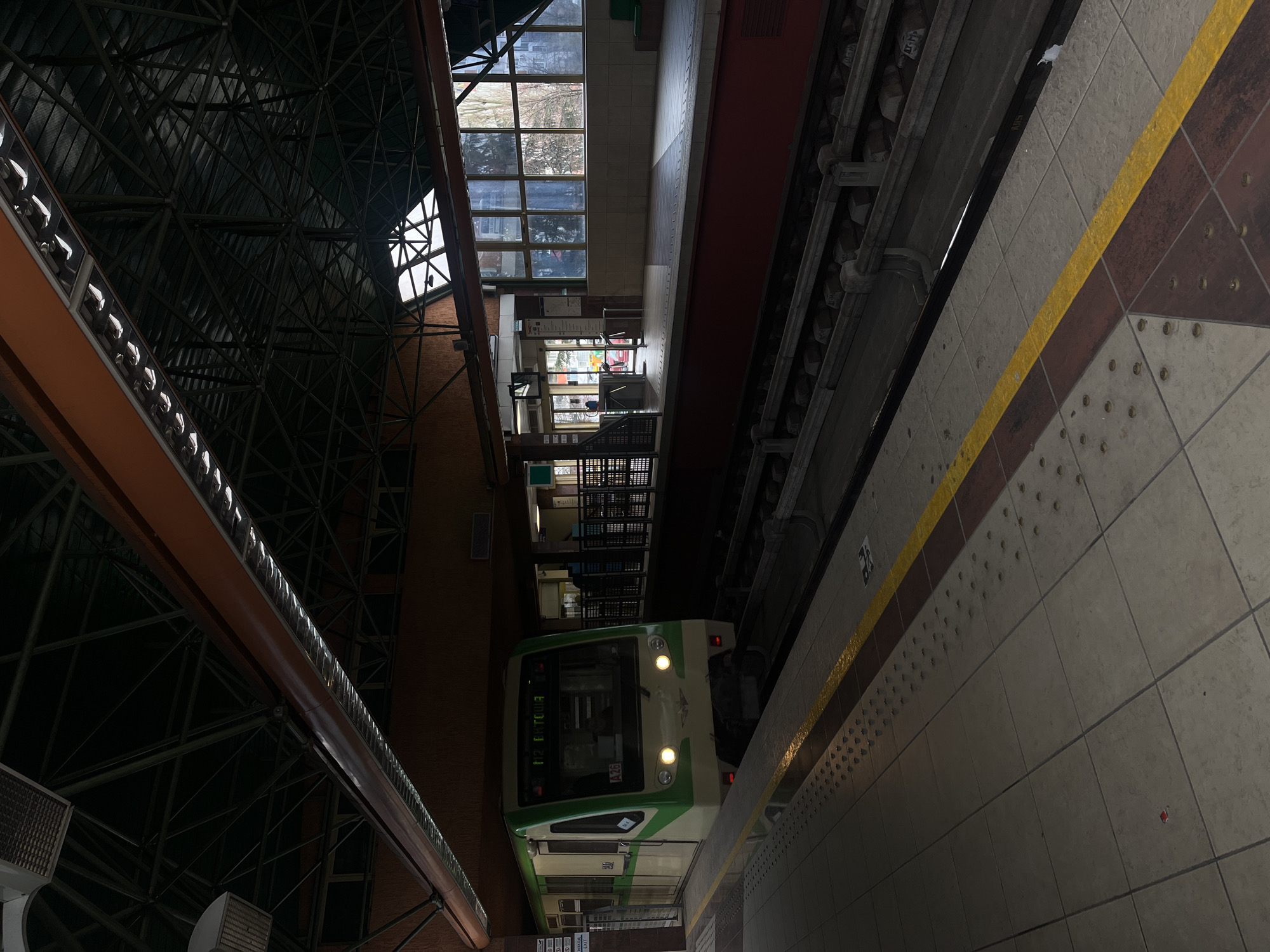 The image captures the interior of Mladost 1 metro station in Sofia, Bulgaria, an elevated platform environment. A two-car Sofia Metro train, predominantly green and cream with "A26" visible on its front, is stationary at the left platform, facing right. Its front headlights are illuminated, and the destination sign reads "ЛЮЛИН". The platform surface is tiled with light and dark square patterns, featuring a yellow tactile paving strip and an adjacent solid yellow line along the track edge.
The station ceiling comprises a dark, exposed metal truss structure with long fluorescent lighting fixtures mounted on orange horizontal beams. A skylight, also featuring a similar truss pattern, allows natural light to enter from above. Large, multi-pane windows extend along the right side of the station, offering an external view of bare trees and residential buildings, suggesting an outdoor or partially uncovered setting.
On the opposite platform, a red bench is visible. Beyond this platform, turnstiles and glass exit doors lead to an exterior area where a red vehicle is partially visible. A single individual is discernible standing in the distance on the far side of the station, beyond the opposite platform and railings. An "ИЗХОД / EXIT" sign in red and white is affixed to a pillar near the train on the left platform. The scene is illuminated by a combination of artificial overhead lighting and natural light from the windows and skylight, resulting in a moderately dim overall ambient light. No direct interactions or significant activity are observable.