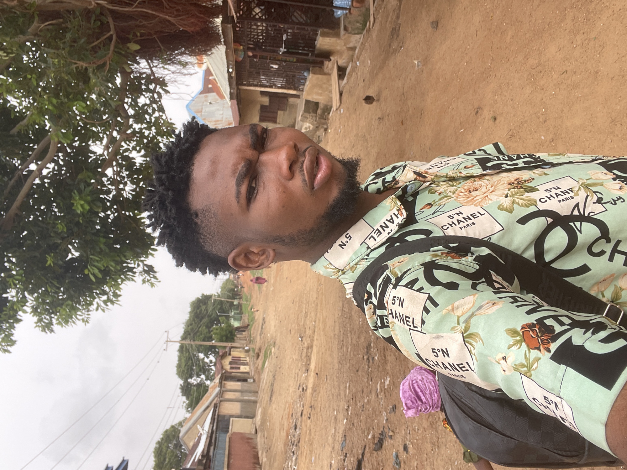 A young man stands in Unguwar Gabas, Nigeria, wearing a light green shirt patterned with Chanel logos and floral designs. He has short, dark hair and a trimmed beard. He is facing the camera with a slight frown. A black bag is slung over his shoulder.
The background shows a dirt road with buildings on either side, utility poles, and greenery. The sky is overcast. Another person is present, partially visible behind the man wearing the Chanel shirt; a purple head covering and part of a dark outfit are visible.