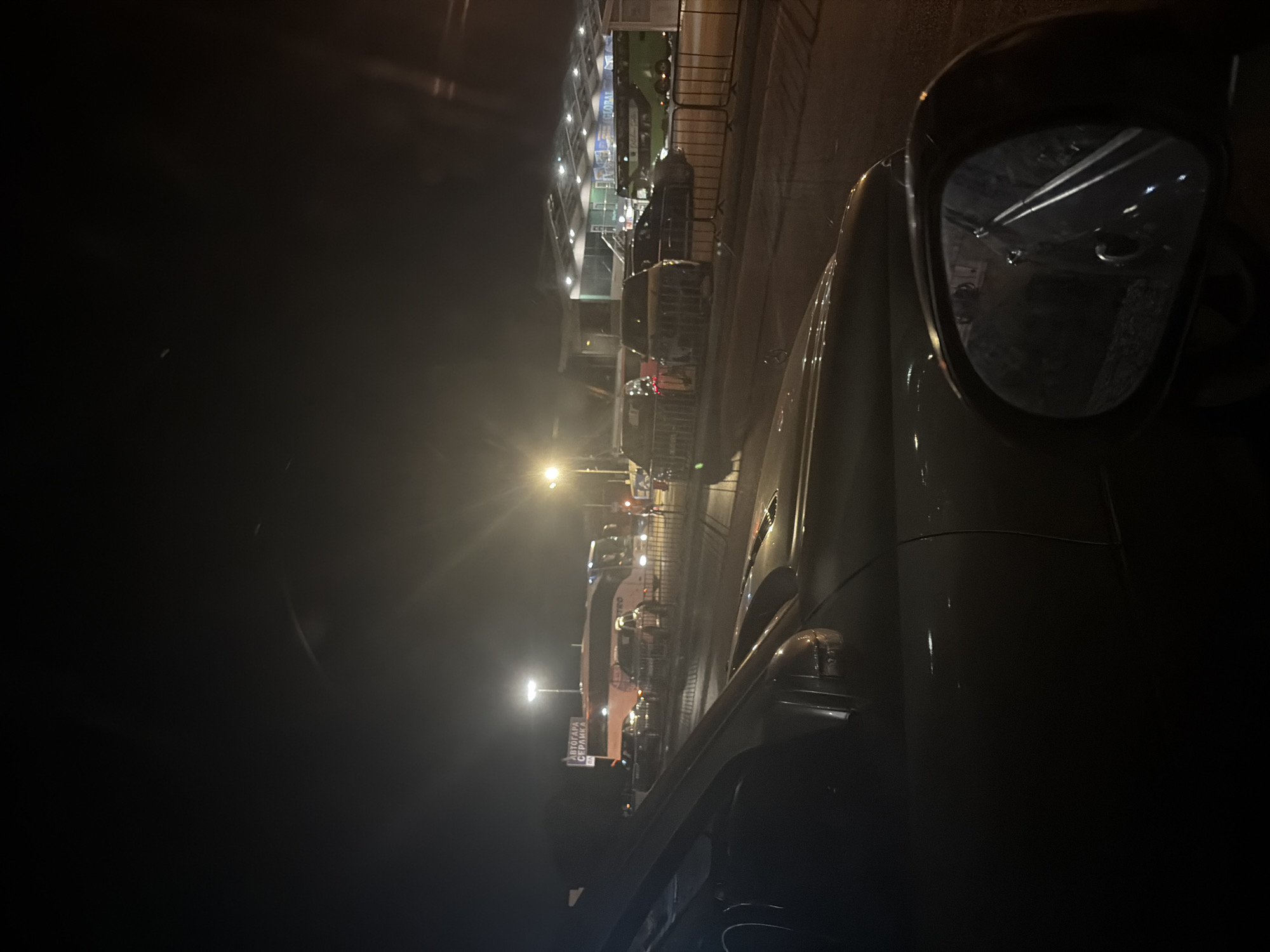 The image depicts a night scene at a bus station or transportation hub, likely in Sofia, Bulgaria. A dark-colored car is in the foreground, seen from the driver's perspective. The car's side mirror is visible, reflecting a dimly lit environment with some railings and structures.
Beyond the car, several vehicles are parked or queuing, including cars and buses. A large white and orange bus with "METRO" visible on its side is prominent. In the background, there's a building with a large, illuminated roof structure, appearing to be a terminal or waiting area. A sign with Cyrillic text, "АВТОГАРА СЕРДИКА" (Avtogara Serdika), is visible on a pole, confirming the location as Serdika Bus Station in Sofia. Streetlights and building lights illuminate the scene, creating reflections on the wet ground. The overall atmosphere is dark and functional, suggesting a typical evening at a transportation hub. No people are clearly visible in the image.