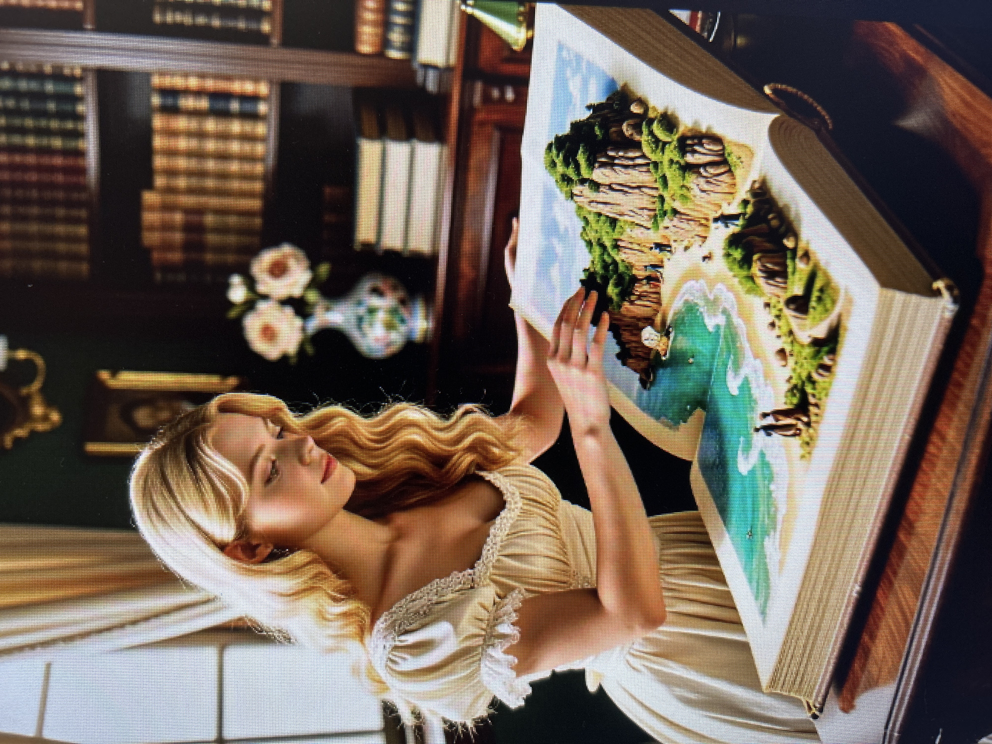 In an indoor setting resembling a library or study, a young woman with blonde, wavy hair is seated at a wooden desk. She is wearing a light-colored, puffed-sleeve dress adorned with pearls and lace detailing around the neckline. Her gaze is directed downwards towards a large, open book placed on the desk in front of her. The book's pages display a vivid, three-dimensional diorama of a coastal landscape, complete with rocky cliffs, lush green vegetation, a sandy beach, and turquoise water. Small figures are visible within the diorama, suggesting a scene of human activity. The background of the scene features bookshelves filled with numerous books, a vase with two light-colored flowers, and a window with draped curtains. A green desk lamp is also partially visible to the right of the book. The overall atmosphere is one of quiet study or imaginative engagement with the book's contents.