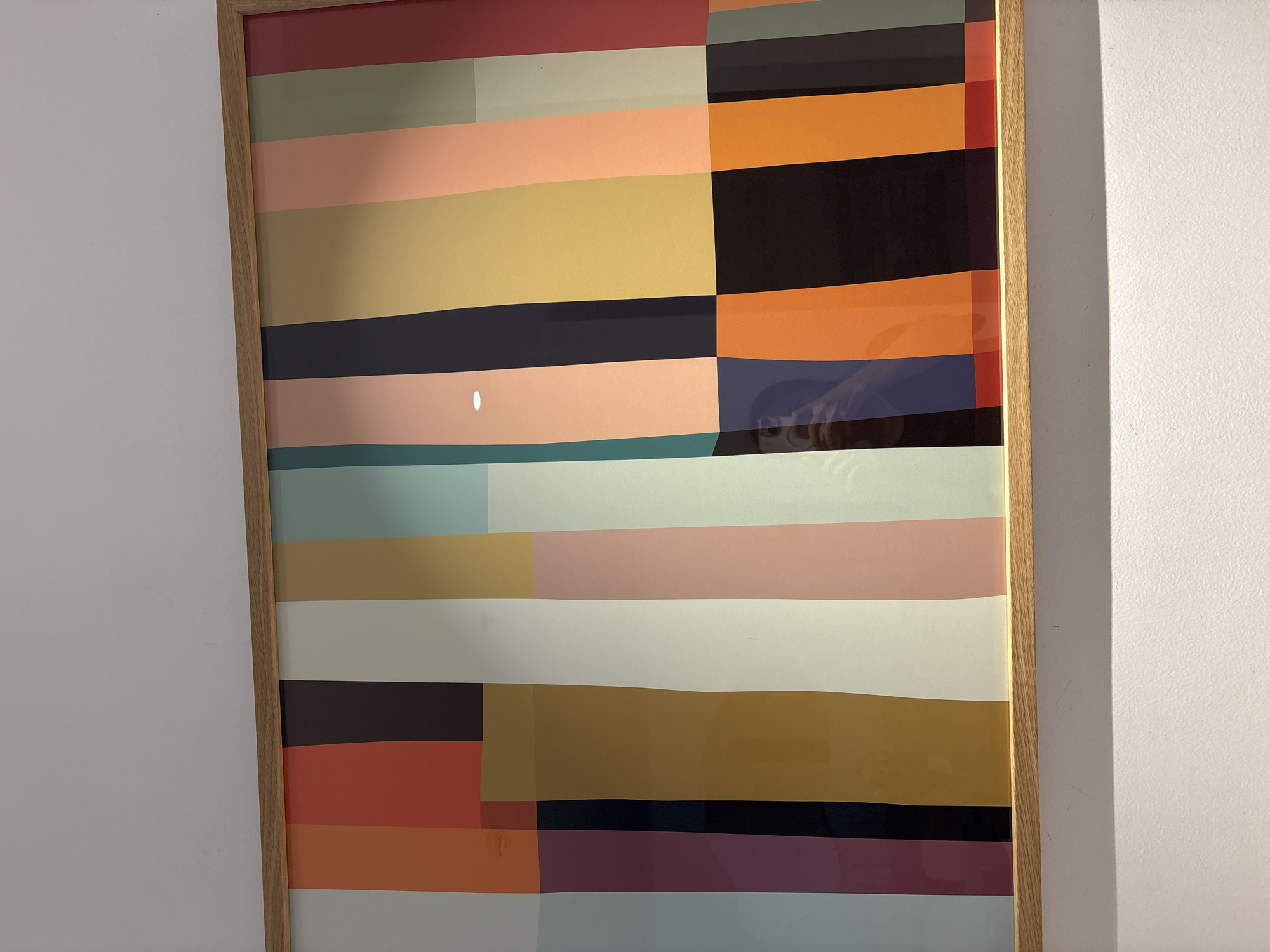 The image shows a framed piece of abstract art with vertical stripes of various colors, including pale gray, orange, red, dark brown, cream, light brown, teal, pink, navy blue, yellow, peach, and dark green. The artwork is framed in a light-colored wood. The artwork is hanging on a white wall, and a shadow is cast on the wall below it. The reflection of a person taking the photo is visible in the artwork. The location is not specified in the image, but it is described as being in Dubai, United Arab Emirates.