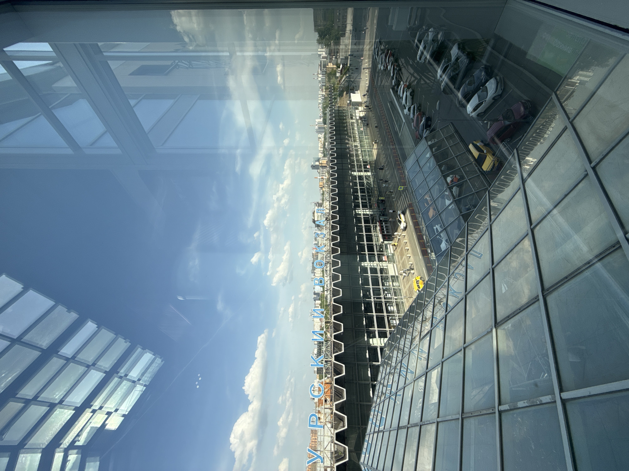 The image shows a cityscape view from within a building with a grid-like window structure. Reflections of the building's interior ceiling are visible on the left side of the frame. Outside, a row of parked cars and a road run parallel to the building. The city extends into the distance, with buildings and greenery visible. A sign with the inscription "Уральский Вокзал" (Uralsky Vokzal, which translates to Uralsky Railway Station) is displayed prominently on a building. This indicates the location is likely in Moscow, Russia.