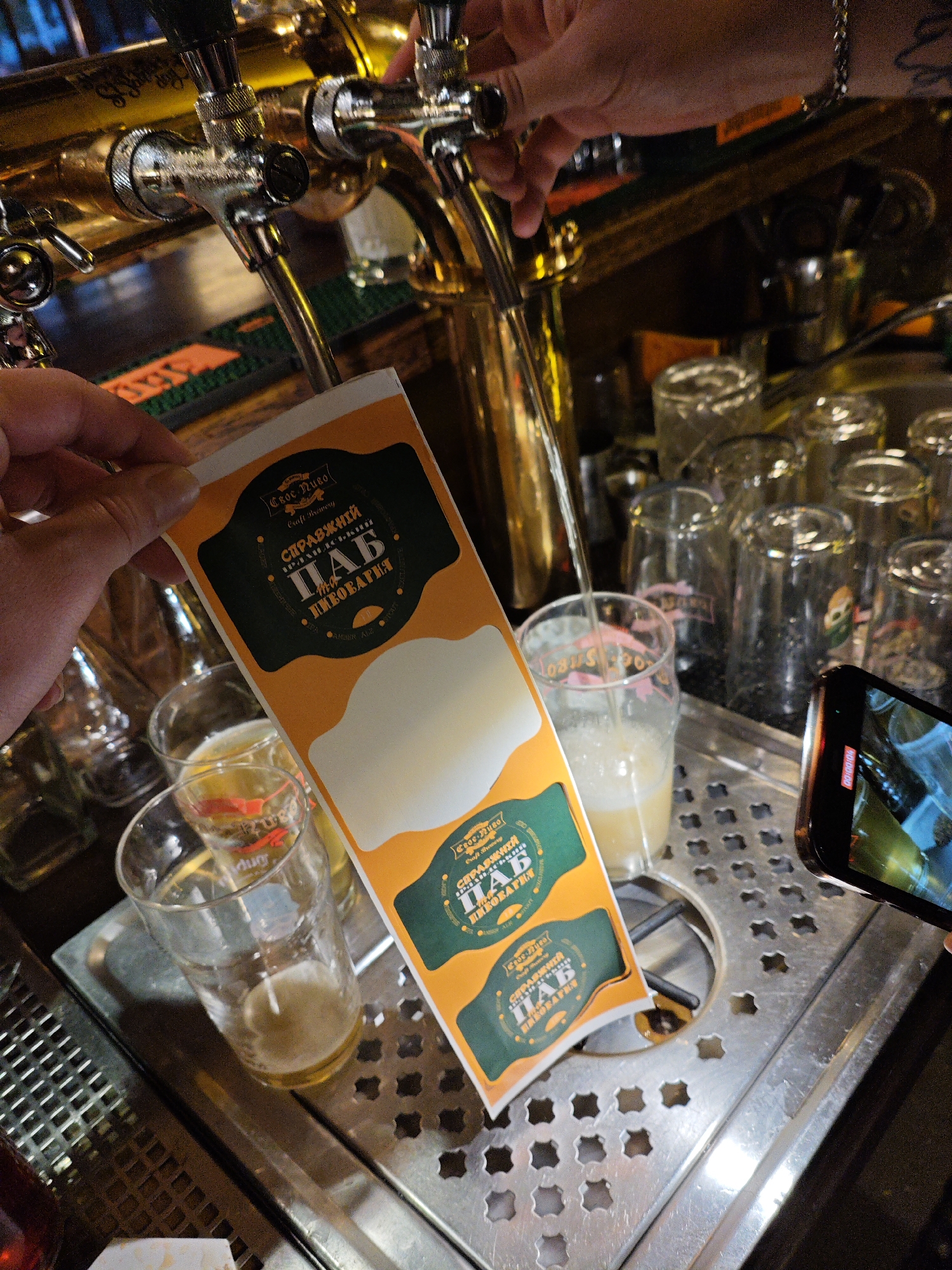 In Kyiv, Ukraine, at a bar or brewery, a hand is holding a beverage label over a glass. The label is orange and green with Cyrillic text that reads "СПРАВЖНІЙ ПАБ" and "ПИВОВАРНЯ." The label appears to be for a craft brewery. In the background, beer taps are visible, along with several empty glasses. A phone is also present, recording the scene. The environment suggests a casual setting, possibly a pub or a bar.