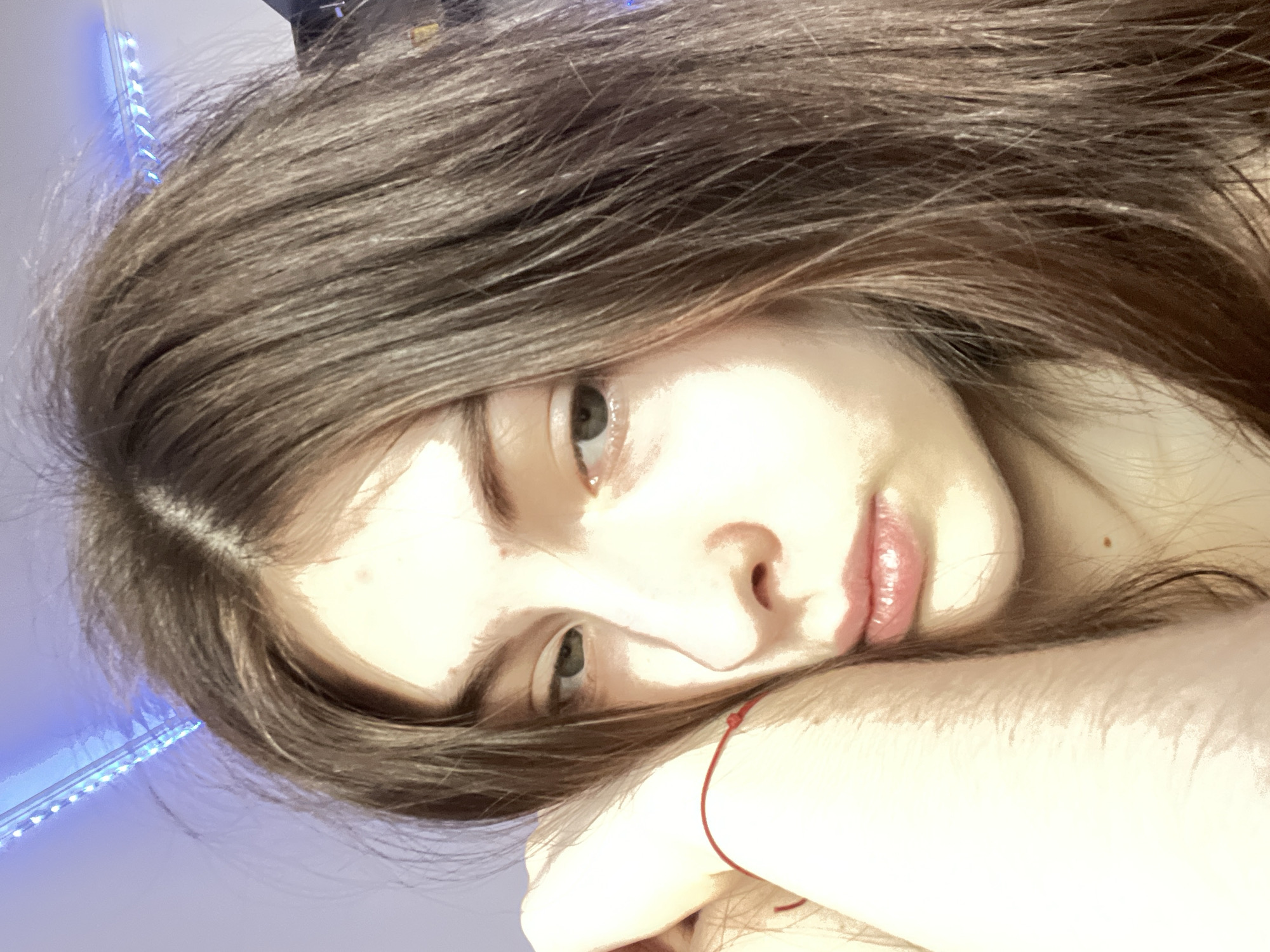 A young female individual is depicted in an indoor setting within the city of Saint Petersburg, Russia. She has long, straight brown hair with a central part, falling over her shoulders and partially obscuring the right side of her face. Her fair skin is significantly illuminated on the right side of her face, with highlights visible on her forehead, cheekbone, and nose, while the left side is in comparative shadow. She possesses light-colored eyes, appearing green or hazel, and full, naturally colored lips. A small, dark brown mole is present on her lower right jawline/neck area. Her left arm is bent, with the forearm supporting her head, and a thin red string bracelet is visible on her left wrist. The individual's gaze is directed slightly downwards and to her left, conveying a subdued or introspective expression. The background consists of a plain, light-colored wall. A blue LED strip light is affixed to the upper left portion of the wall, casting a distinct blue illumination, with a similar blue light source partially visible in the upper right.