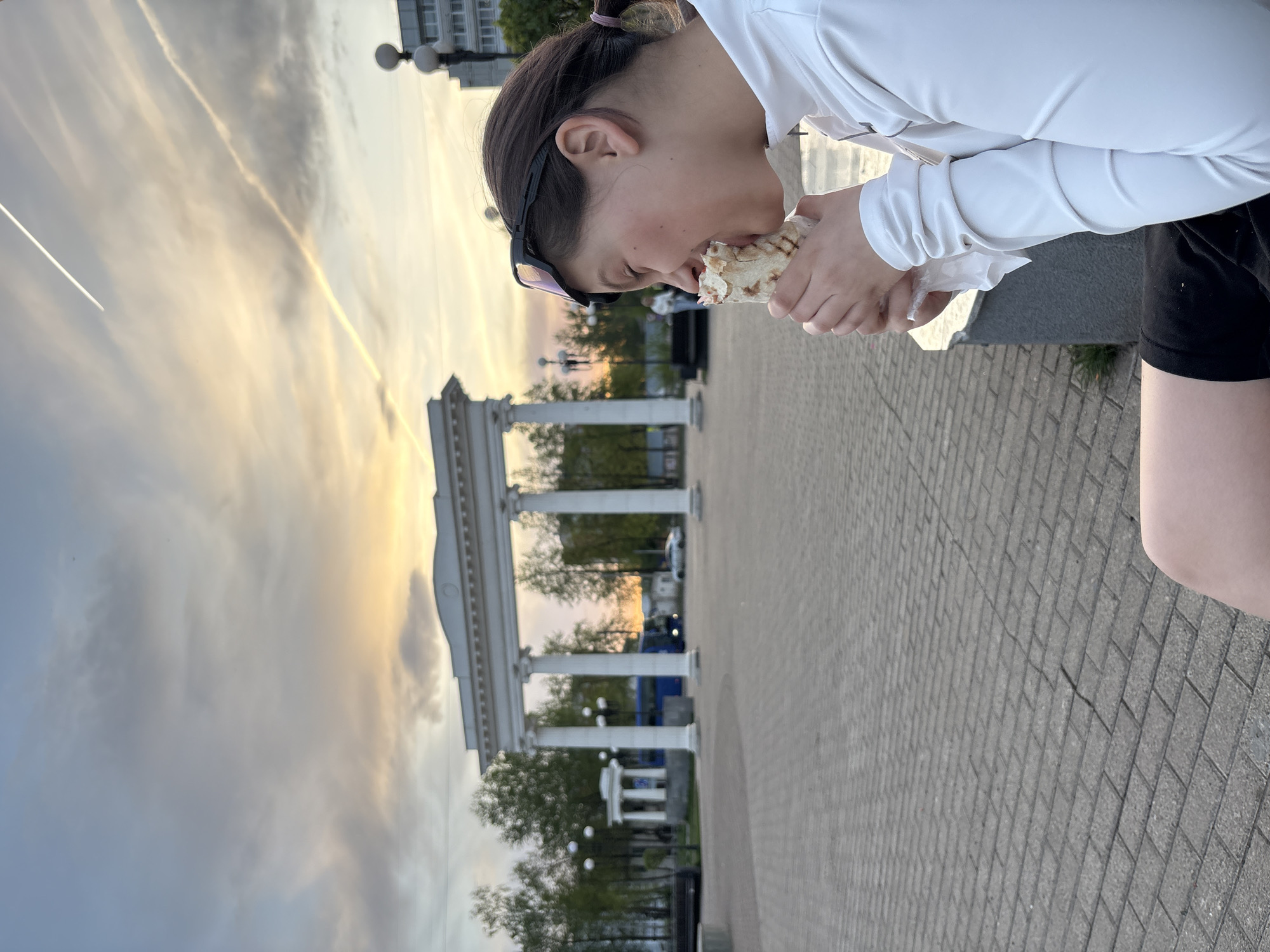 A person with dark hair and wearing a white long-sleeved shirt and black shorts is eating a wrap while standing on a brick-paved area. The person is facing right, and sunglasses are pushed up on their head. In the background is an architectural structure with white columns and arches, identifiable as the colonnade on Karl Marx Street in Chaikovsky, Russia. Behind the colonnade are trees and possibly buildings. The sky is a mix of grey and golden colors, possibly indicating sunset or sunrise.