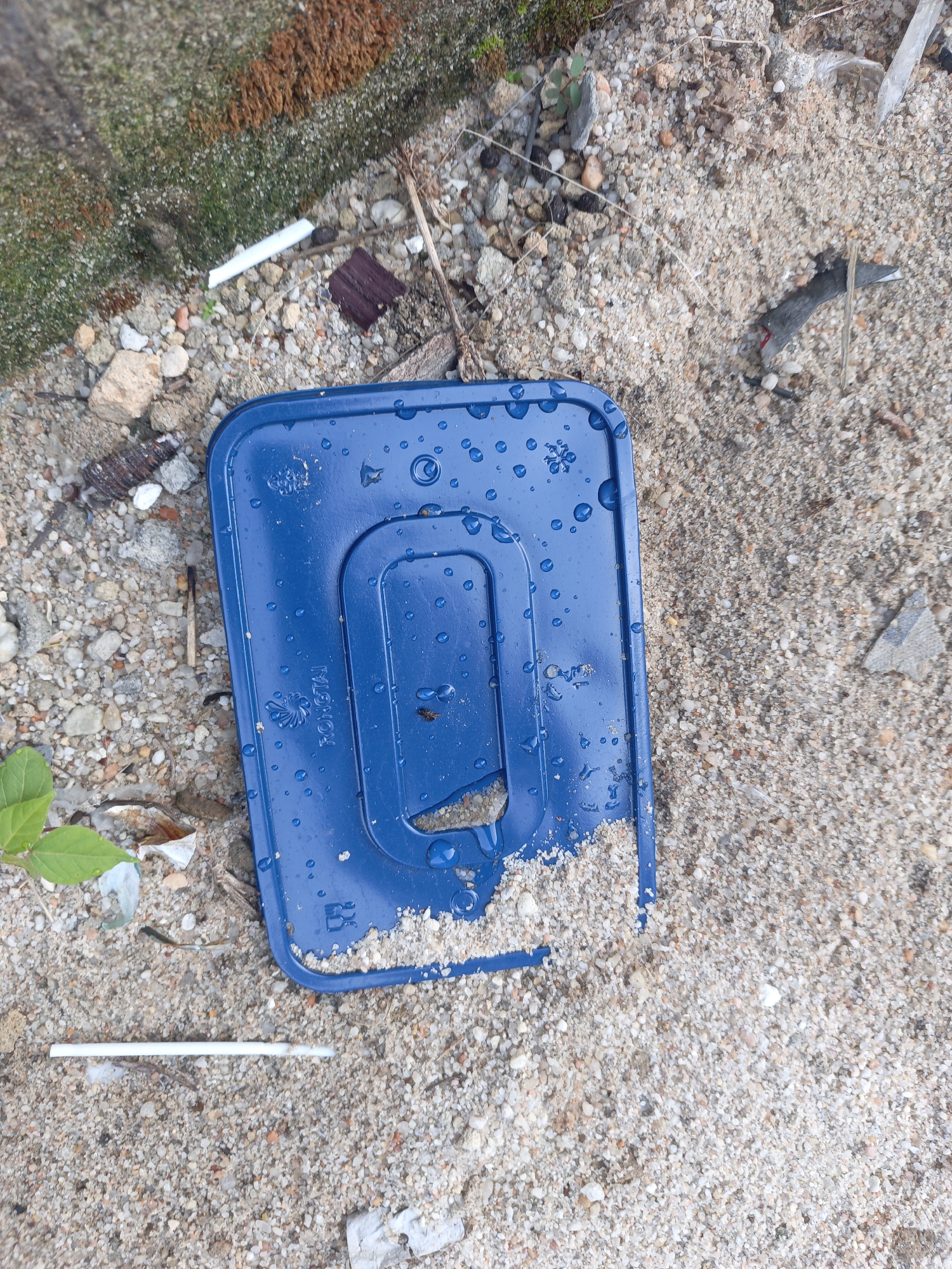 The image shows a discarded, rectangular, blue plastic object with rounded corners lying on sand mixed with small pebbles. Water droplets are scattered across the surface of the plastic. The brand "RONGTAI" is imprinted on one side. The object is damaged with a broken section on one edge. Scattered around the plastic are bits of debris, including cigarette butts, small pieces of wood, and a few green leaves. Moss grows on the top left, next to concrete. The location is a patch of waste ground.