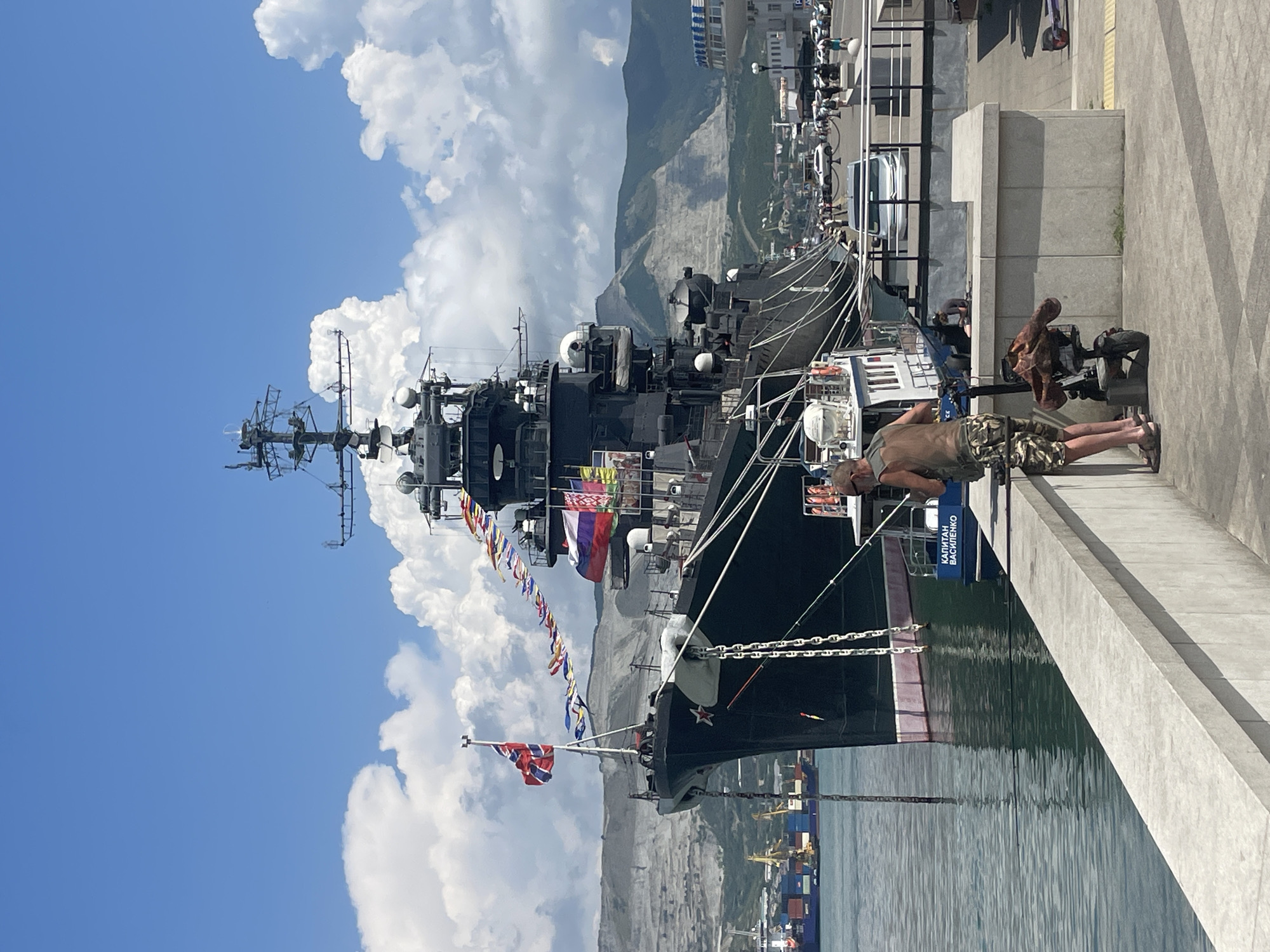 A large, dark-colored warship, identified by a red star insignia and Russian flags, is docked in the port of Novorossiysk, Russia. The ship flies numerous colorful signal flags along its bow. In the foreground, a man in camouflage shorts stands on a concrete promenade, facing the water and seemingly engaged in fishing with a rod. Beside him is a parked electric scooter with a bag attached. Behind the man, a smaller white boat with the Cyrillic inscription "КАПИТАН ВАСИЛЕПКО" is moored. The background shows a hillside with visible quarry marks, port infrastructure including cranes and container ships, and a clear blue sky with scattered white clouds. A road with cars and pedestrians is visible along the waterfront beyond the promenade.