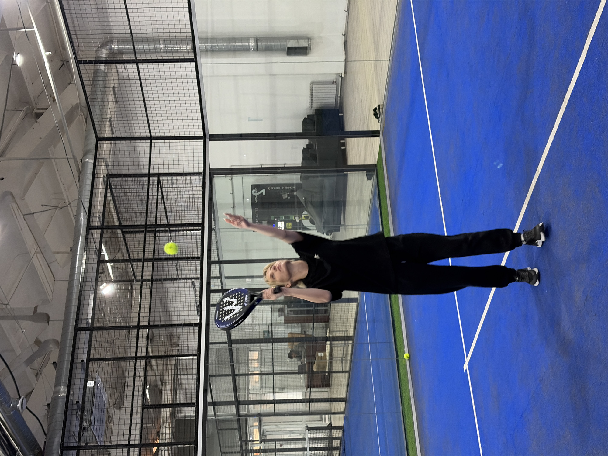 A man is playing padel tennis on a blue court surrounded by a metal cage and glass walls. The man is wearing black clothing and dark sneakers. He is holding a padel racket in his right hand, reaching up to hit a yellow ball. Another yellow ball is on the blue court. The location is likely a padel tennis facility. It is not possible to confirm this is in Chaikovsky, Russia based on the image.