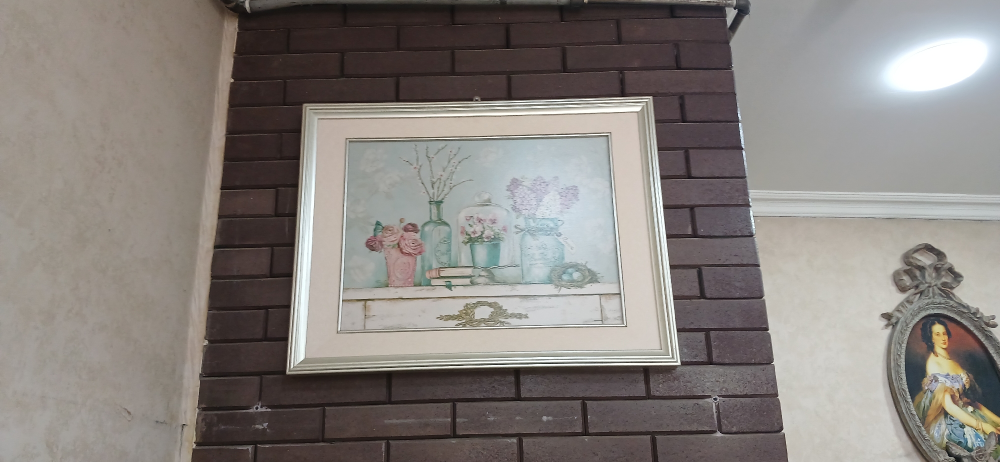 The image shows a framed picture of flowers and decorative objects on a mantelpiece, hanging on a brick wall. To the right of the brick wall, a decorative oval frame with a painting of a woman in historical attire is visible. The environment appears to be indoors, possibly in a home or a museum. No specific location in Ganja City, Azerbaijan can be determined from the image.
