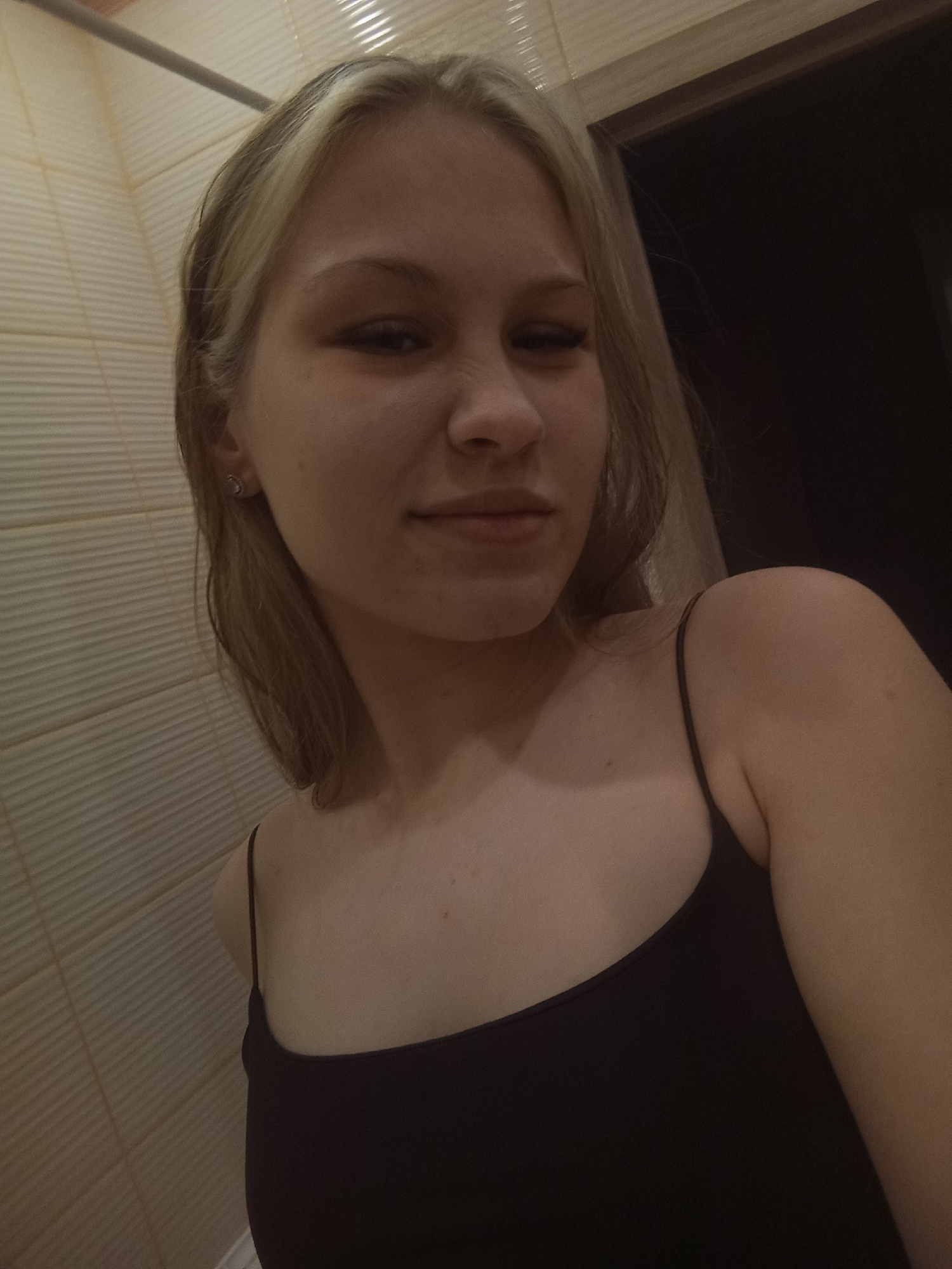 A young woman with blonde hair and a black tank top is in a bathroom. The bathroom has light-colored tiled walls. There is a doorframe visible in the background. The location is not discernible as Krasnodar, Russia based on the provided image.