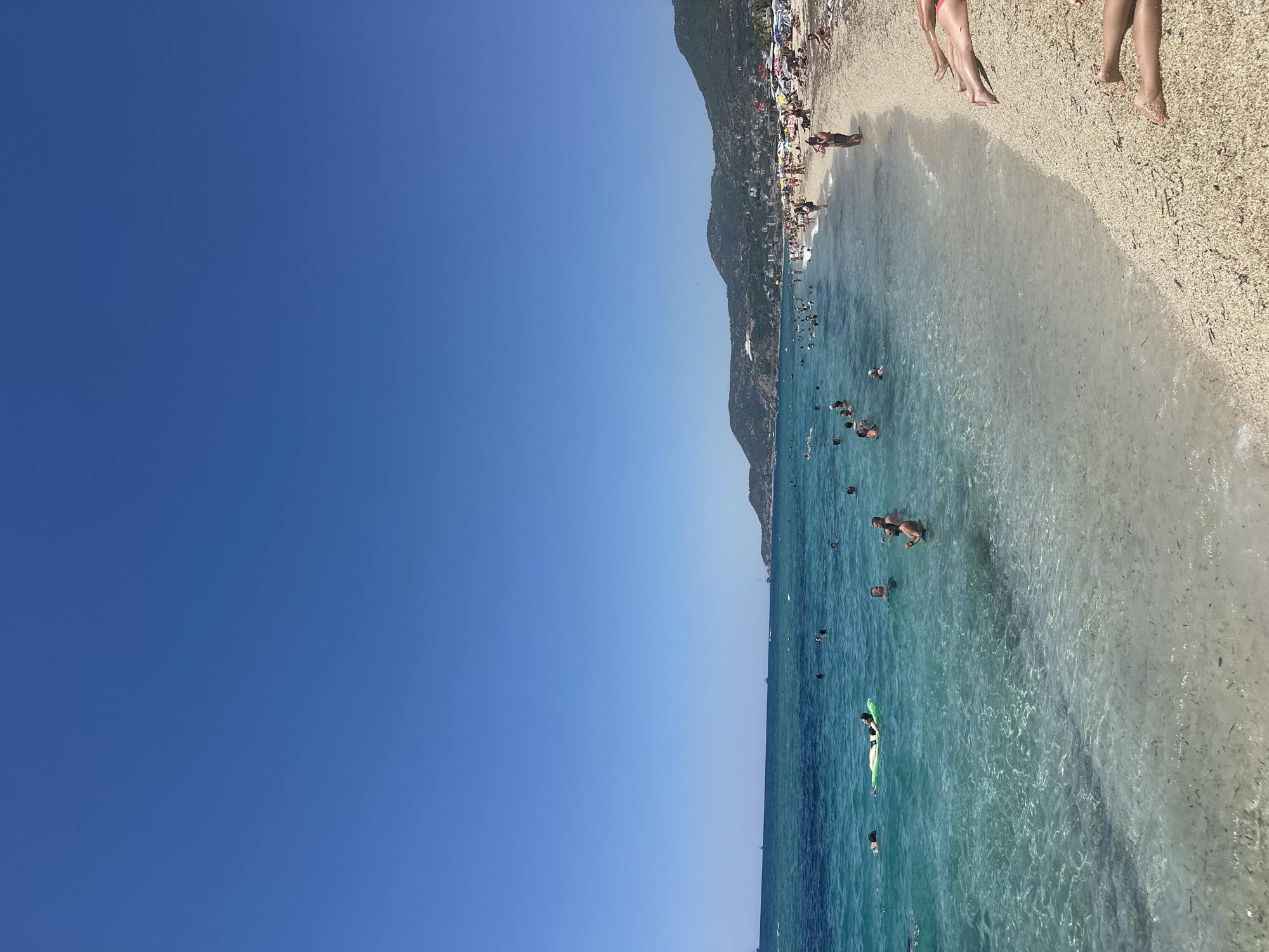 This is a long-shot, eye-level, outdoor, daytime photograph of a crowded beach scene in Alanya, Turkey. The image captures a clear, bright blue sky dominating the upper portion, transitioning to a turquoise, clear ocean with many people swimming and floating on inflatables. The water is shallow near the shore, with visible sand and small debris beneath the surface. A sandy and pebbly beach lines the right side of the frame, filled with sunbathers relaxing on towels and under umbrellas. The background reveals a hilly, green landscape with buildings and more umbrellas scattered along the coastline, indicating a populated resort area. Several boats are visible in the distance on the sea. The overall atmosphere is one of leisure and summer vacation activities.