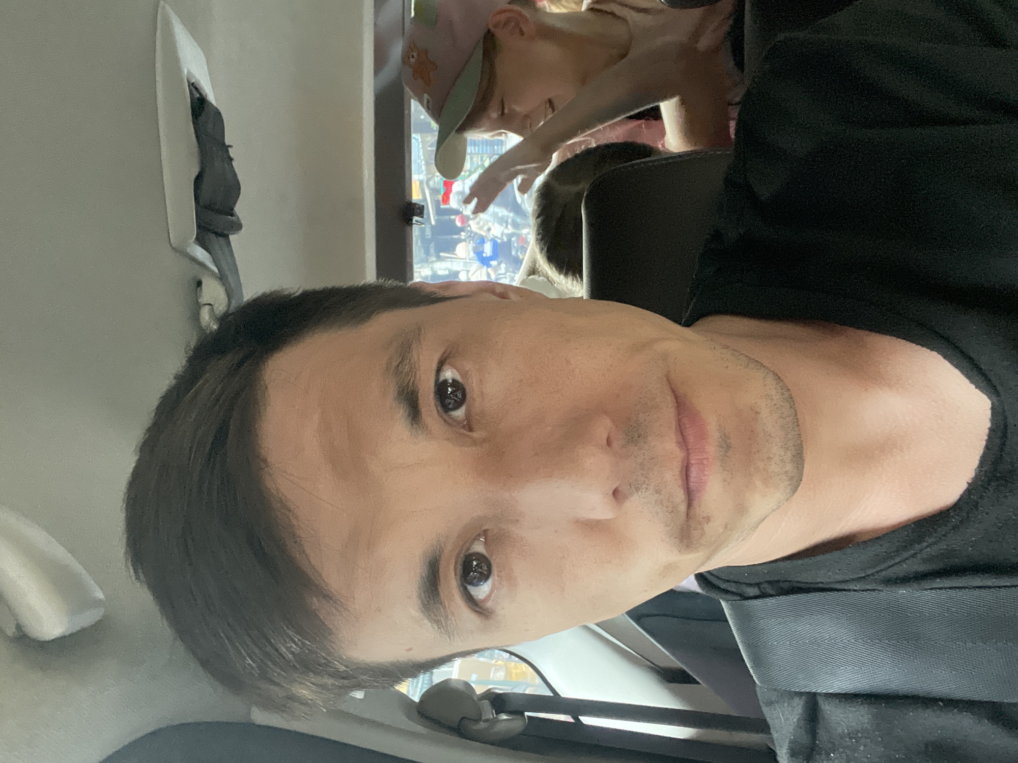 This is a selfie taken from inside a vehicle, likely a car or van, by a man in the foreground. He is wearing a black t-shirt and a seatbelt is visible across his chest. His gaze is directed at the camera. In the background, through the vehicle's window, a busy city street scene is visible, with motorbikes and pedestrians. There is also a young girl in the back seat of the vehicle, wearing a pink cap and looking to her right, with her hand raised. The lighting suggests it is daytime. The scene is consistent with a location in Thủ Đức, Vietnam, given the presence of motorbikes and the urban environment.