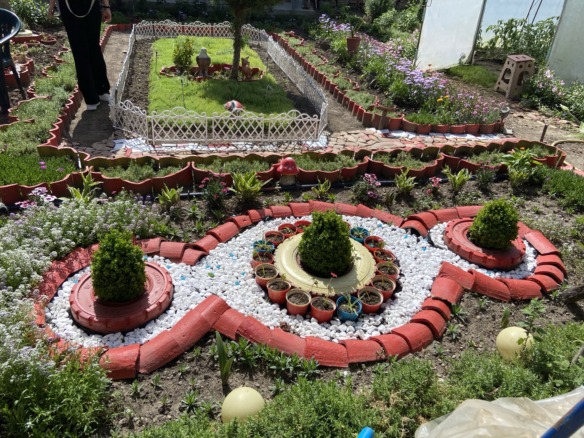 The image depicts a well-tended garden in Djulunitsa, Bulgaria. The garden features elaborate landscaping with intricate designs created using recycled materials, such as painted tires and repurposed flower pots. There are several distinct garden beds separated by pathways made of red clay bricks. Each bed contains a variety of plants and flowers in full bloom. In the background, there is a small patch of green grass contained by a white ornamental fence. A person is seen partially in the image, wearing black pants and white shoes, seemingly walking through the garden. There are a few other objects in the background that appear to be garden tools and accessories. The environment is bright and sunny, suggesting it is daytime.