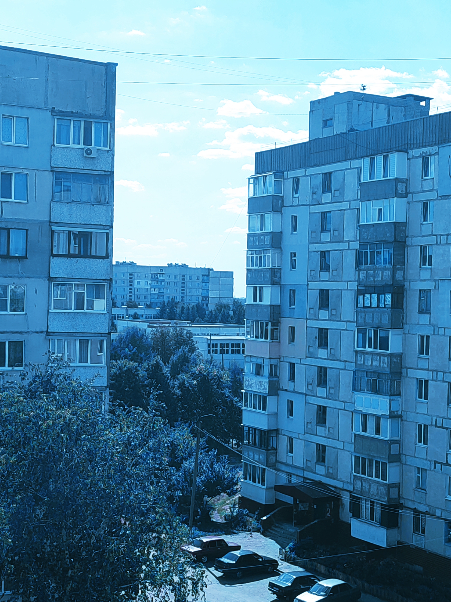 The image depicts a scene in Zlatopil, Ukraine, with a focus on residential buildings and surrounding elements. In the foreground, two multi-story apartment buildings are prominently featured. The building on the left is a light blue, concrete structure with numerous windows and balconies. The building on the right, also a concrete structure, is taller and exhibits a similar architectural style with many windows and enclosed balconies.
Between these buildings, trees with dense foliage, primarily appearing dark blue due to the lighting and color grading, are visible. In the background, more apartment buildings of varying heights and designs can be seen, suggesting a densely populated urban or suburban area. Power lines are strung across the sky, and a few scattered clouds are present in the bright blue sky.
At the base of the buildings, parked cars are visible on a paved area that appears to be a street or parking lot. The cars are dark in color and their details are somewhat obscured. A street lamp is also present. The overall atmosphere of the image is calm and static, portraying a typical day in a residential district of Zlatopil. No people are directly visible in the scene.